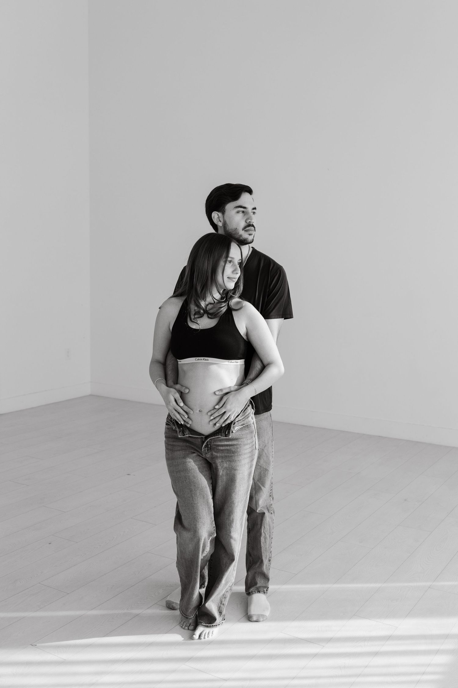 Black and white photo of a man embracing a pregnant woman from behind in a bright room.