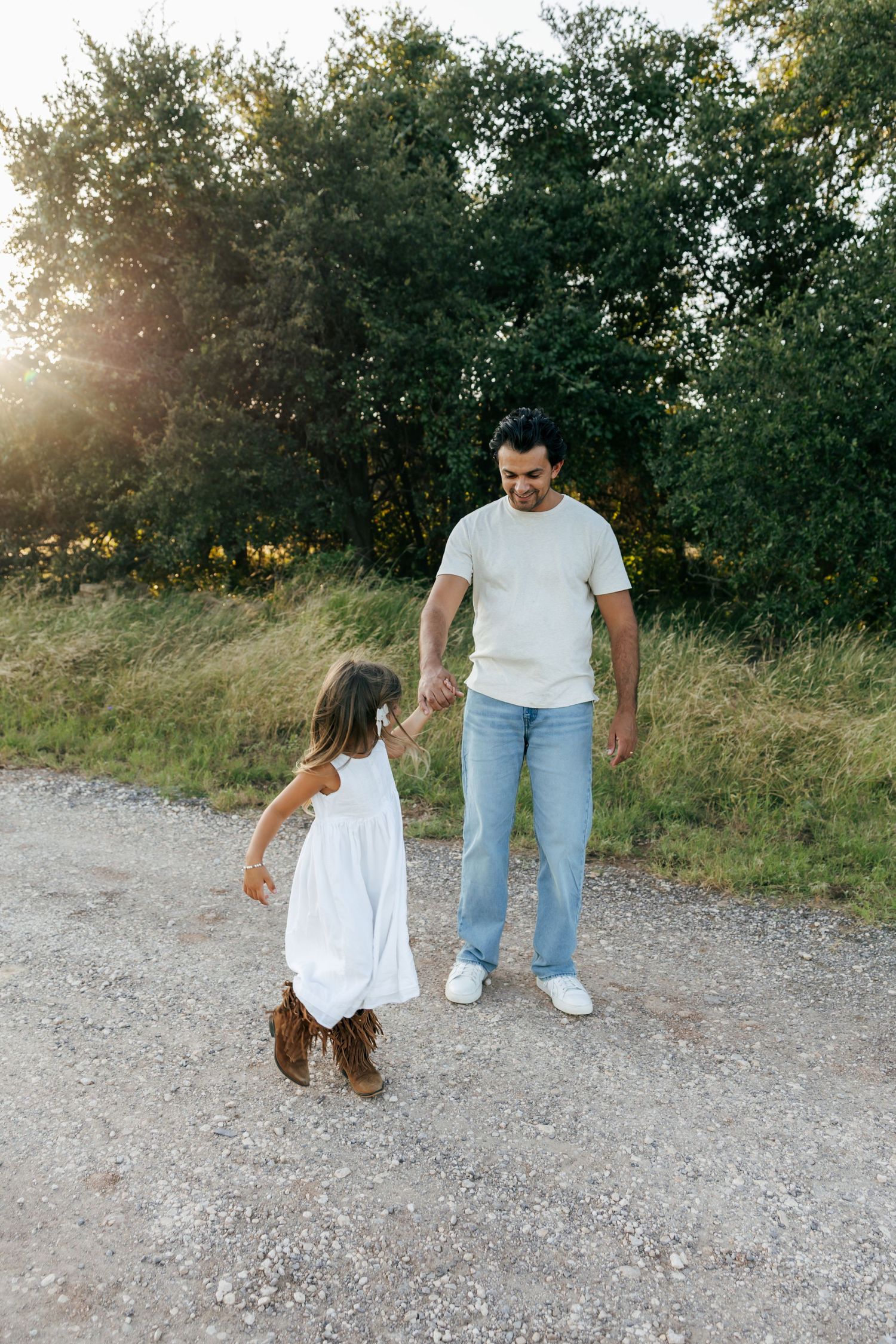 Father and young daughter in white dress holding hands on a gravel path at sunset.