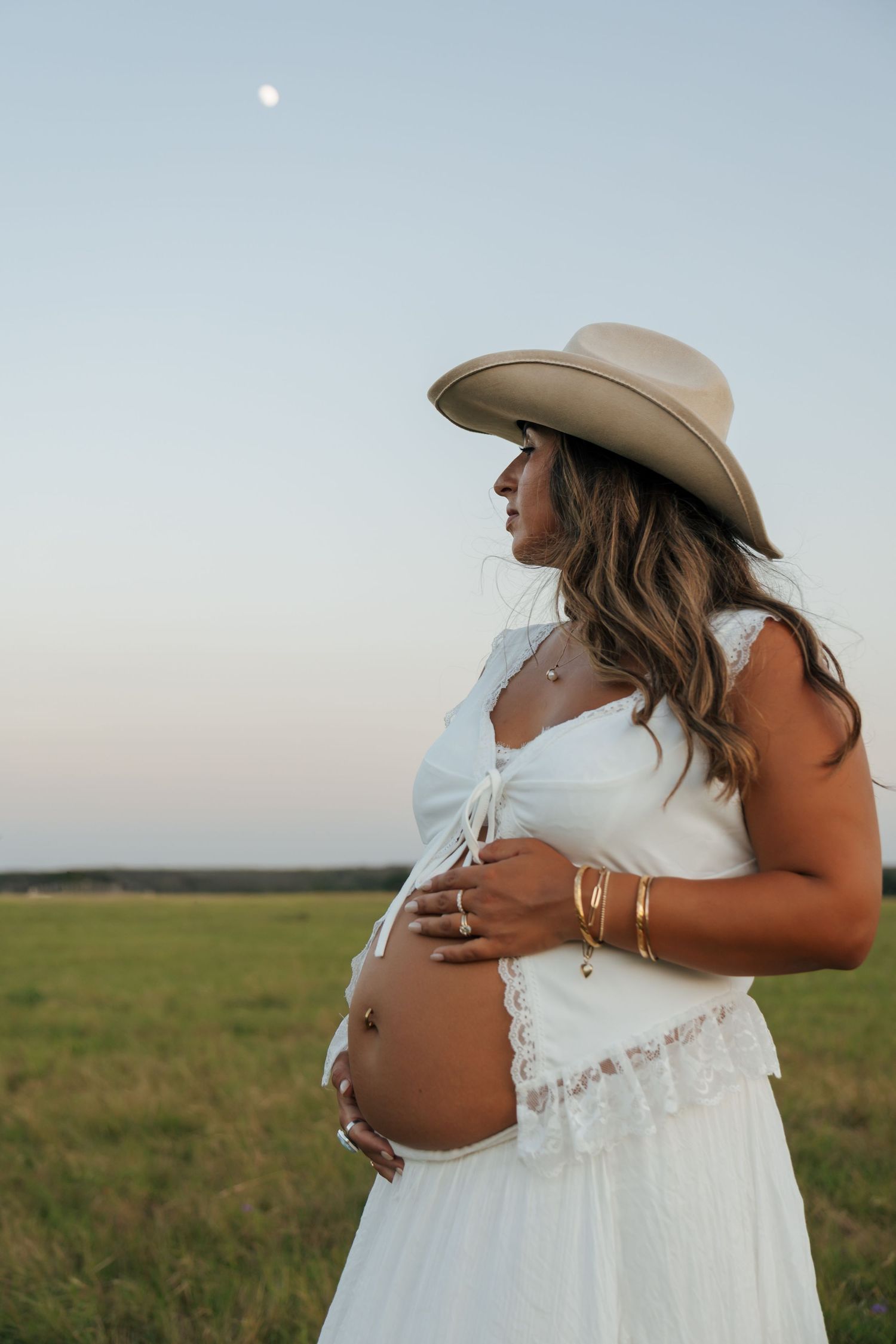 Pregnant woman in white dress and cowboy hat holds her baby bump in a field at dusk.