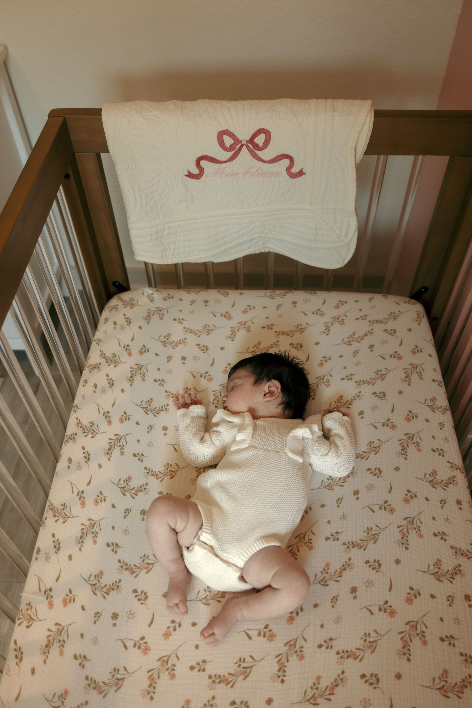 Newborn baby sleeping in wooden crib with floral sheet and decorative bow blanket.