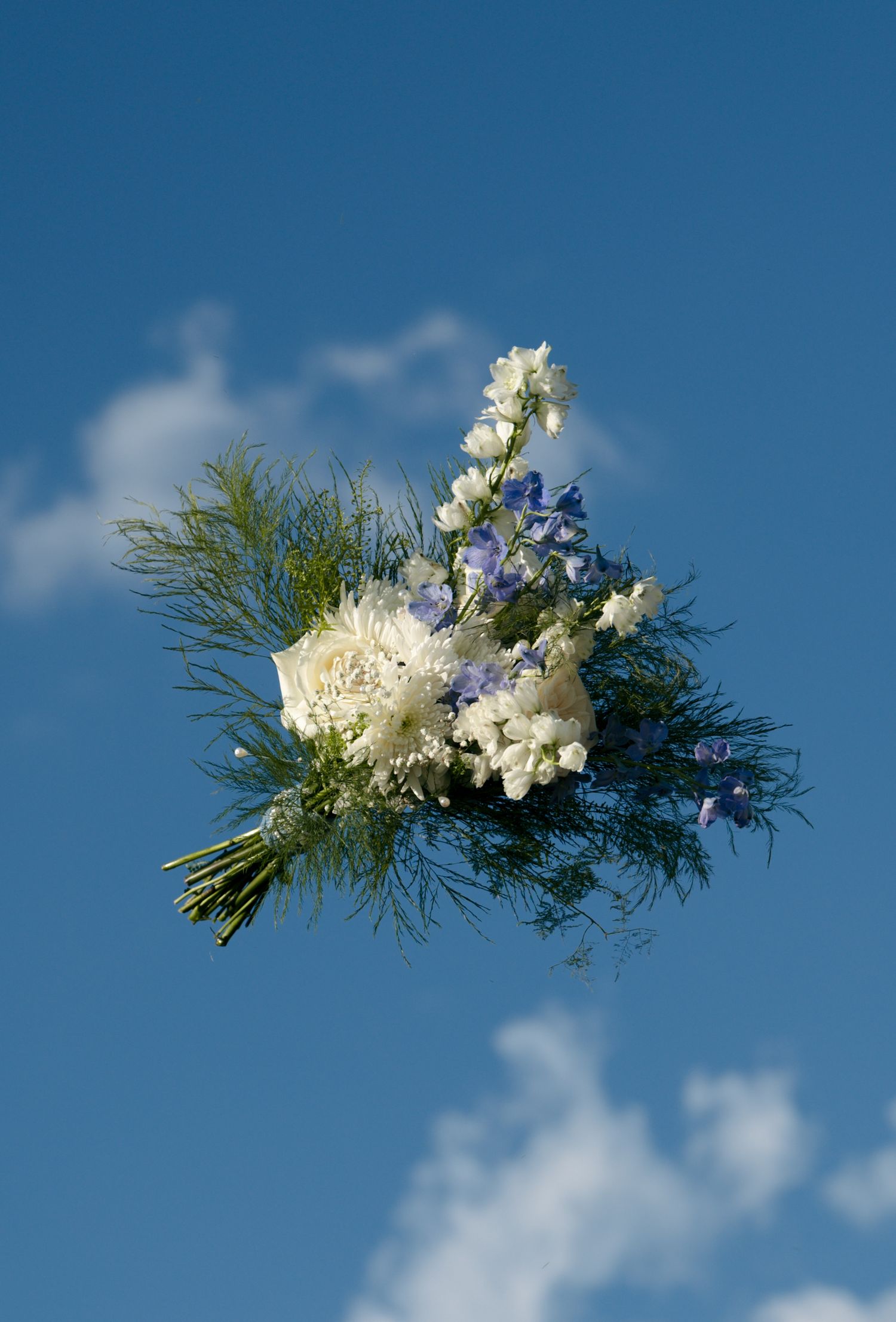 A bridal bouquet of white and blue flowers with green ferns tossed against a bright blue sky.