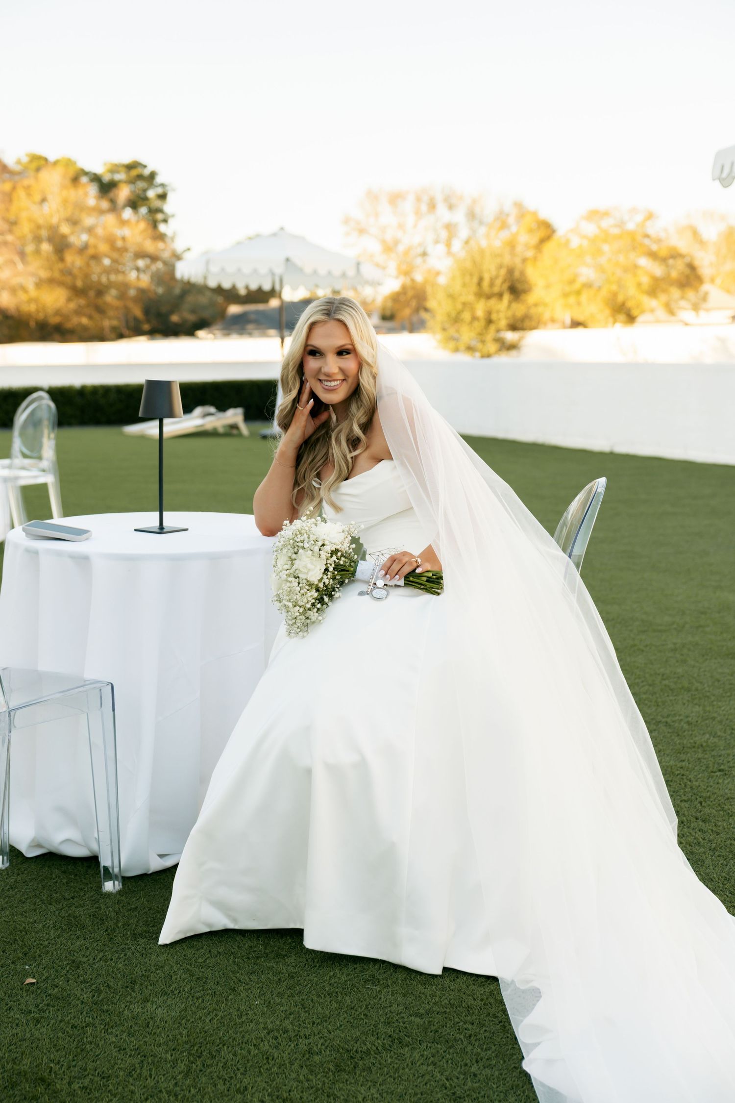 Elegant bride in strapless white gown with veil sitting at outdoor table holding white floral bouquet.