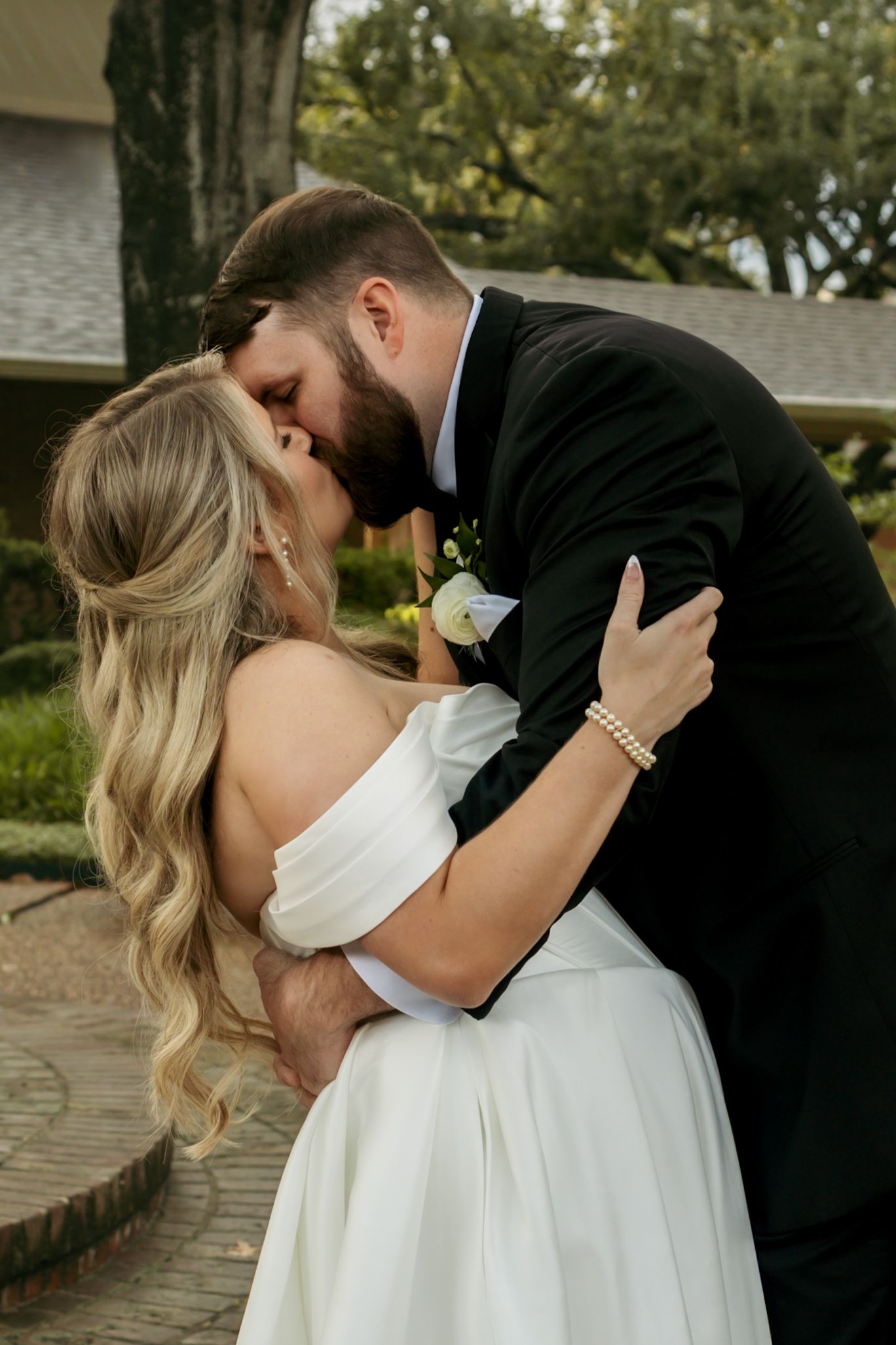 Newlyweds kissing passionately outdoors, bride in off-shoulder white dress with pearl bracelet.