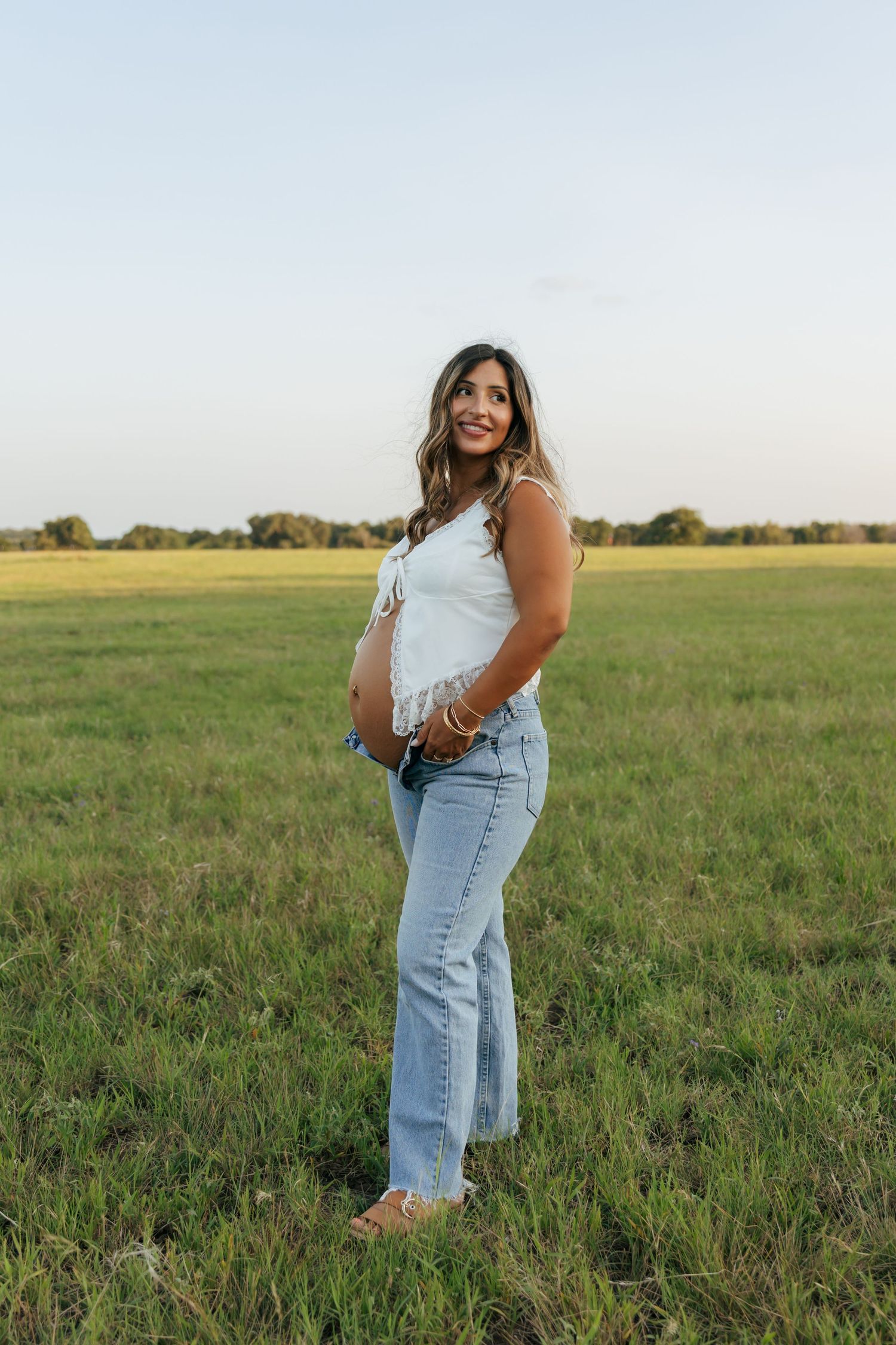 Smiling pregnant woman in white top and jeans stands in an open green field at golden hour.