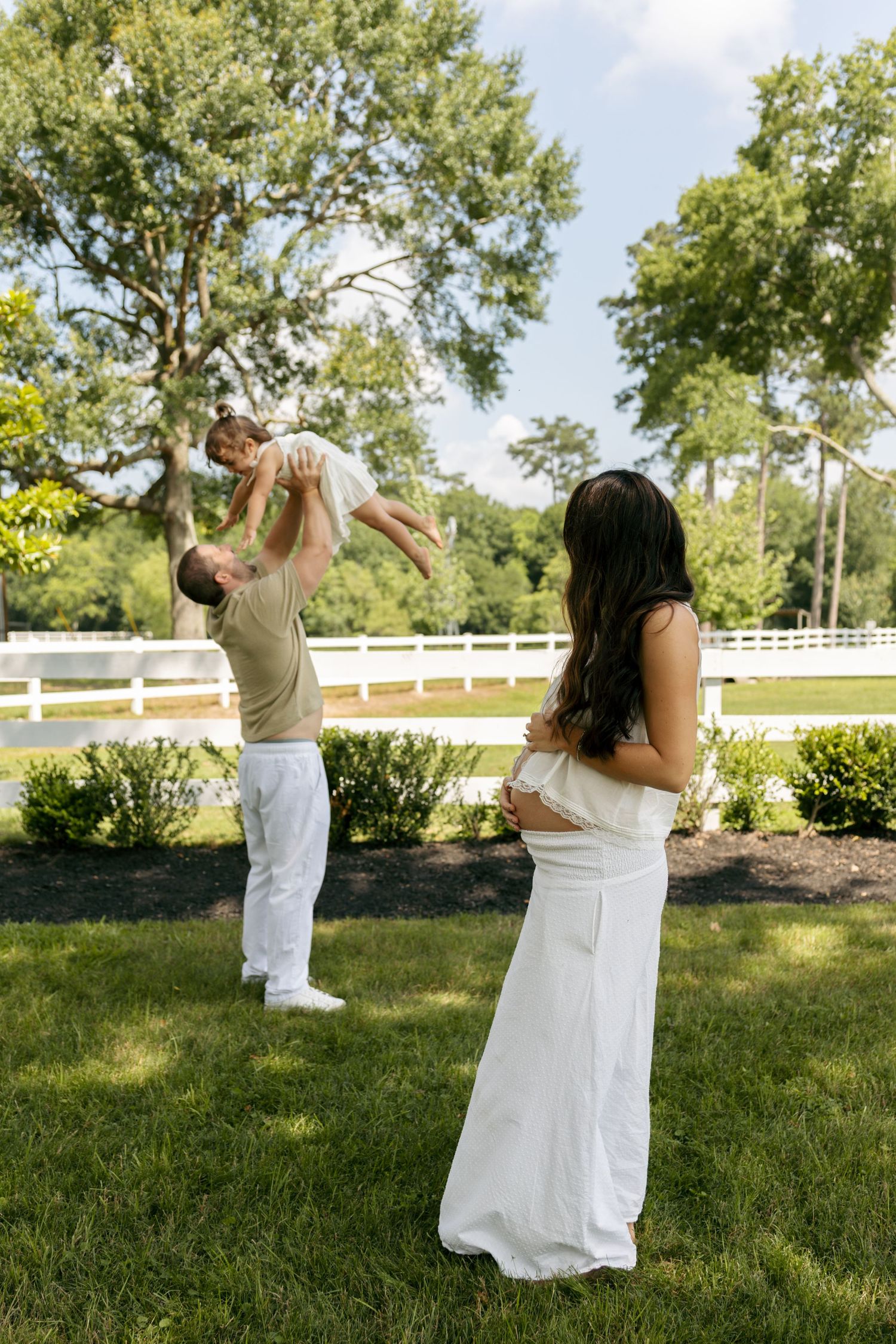 Pregnant woman in white watches as man lifts a toddler in the air in a lush backyard.