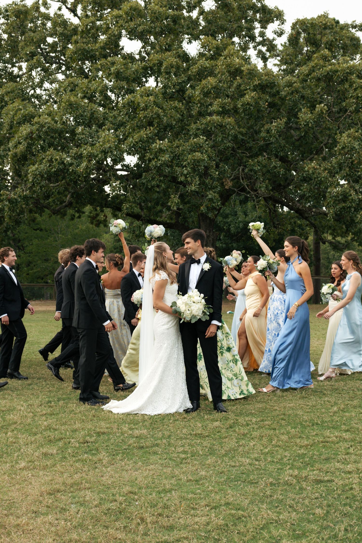 Wedding party celebrating outdoors, bride and groom surrounded by bridesmaids in blue gowns and groomsmen in tuxedos.