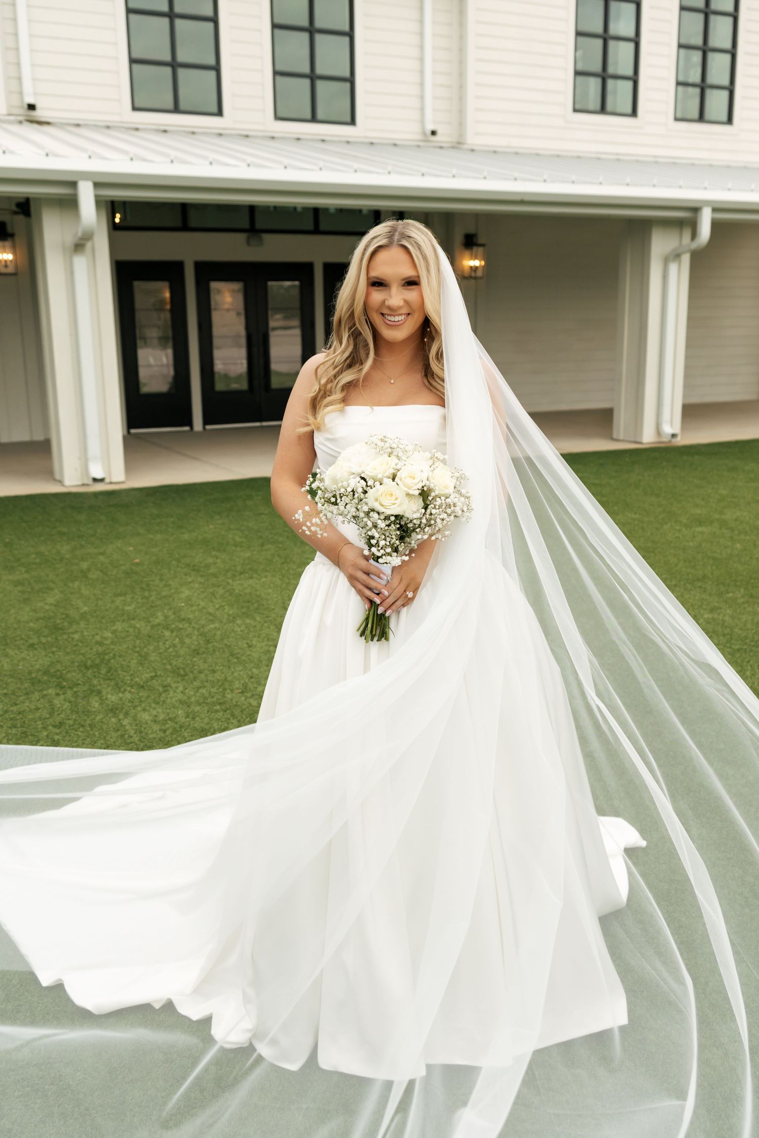 Bride in strapless white ballgown with flowing veil holds white bouquet outdoors at elegant wedding venue.