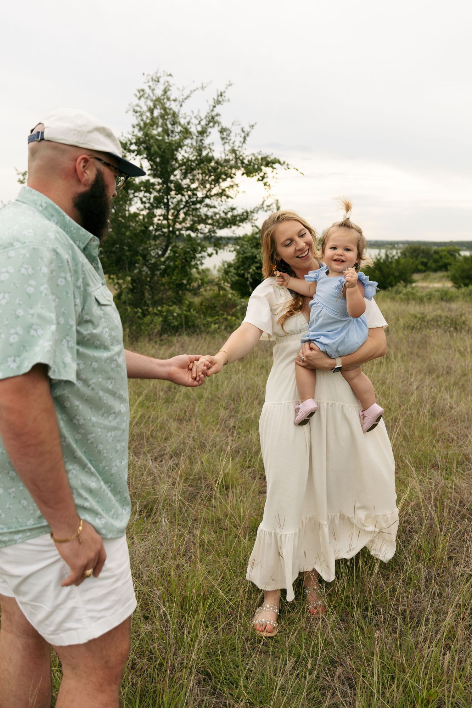 Happy mother in white dress holding toddler while father reaches for her hand in meadow.