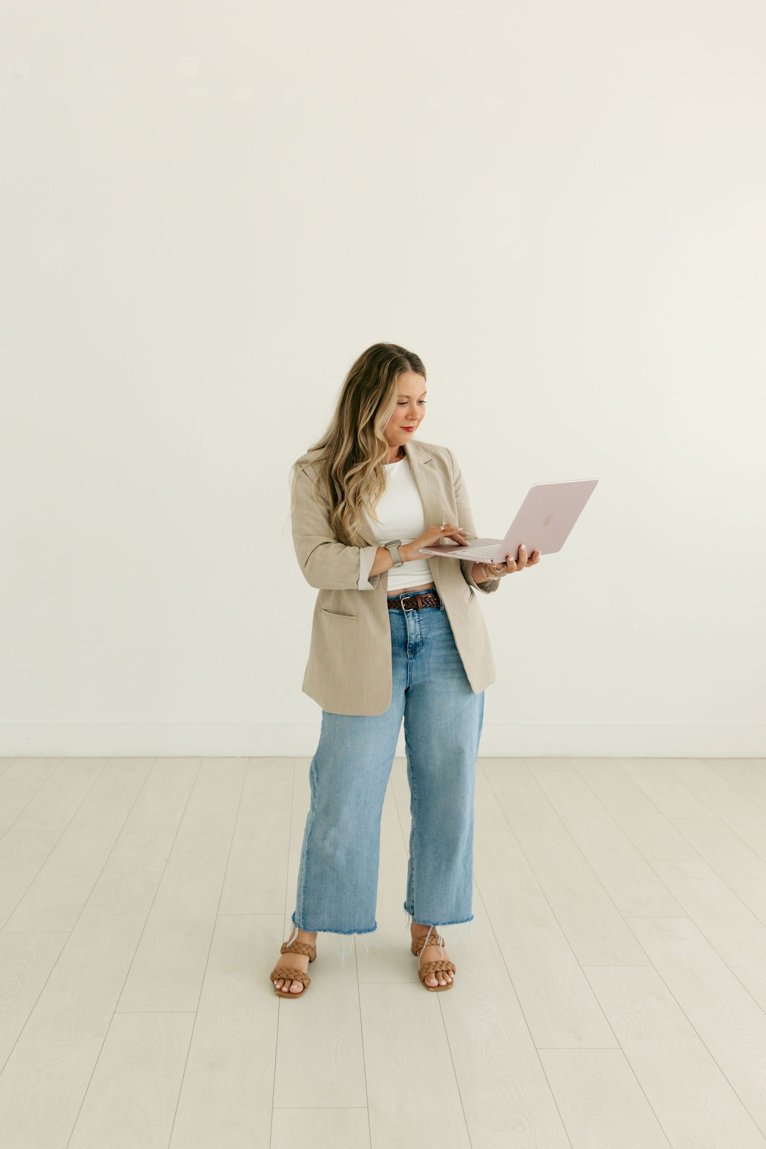 Professional woman in beige blazer and wide-leg jeans standing and working on pink laptop computer.