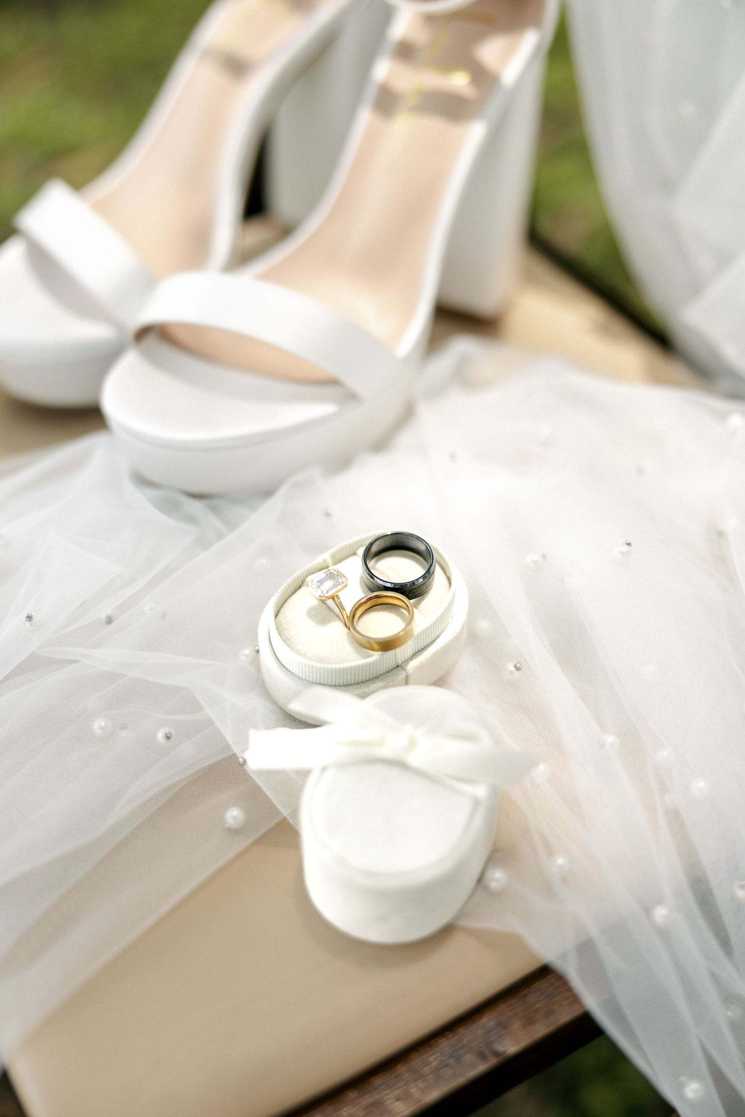 Wedding rings displayed on white ring box beside bridal veil and white platform heels outdoors.