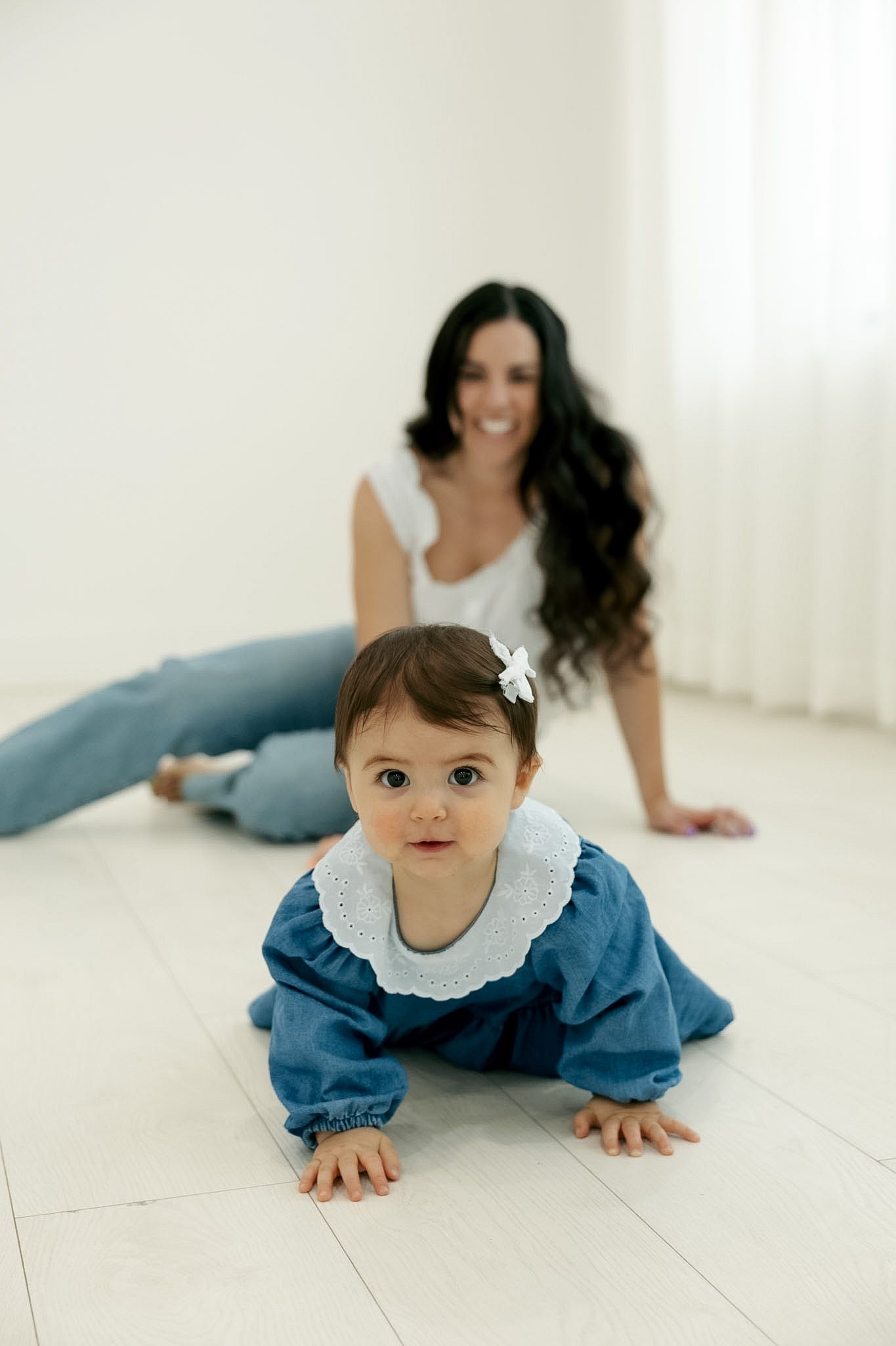 Baby girl in blue dress crawling on white floor while smiling mother watches from behind.