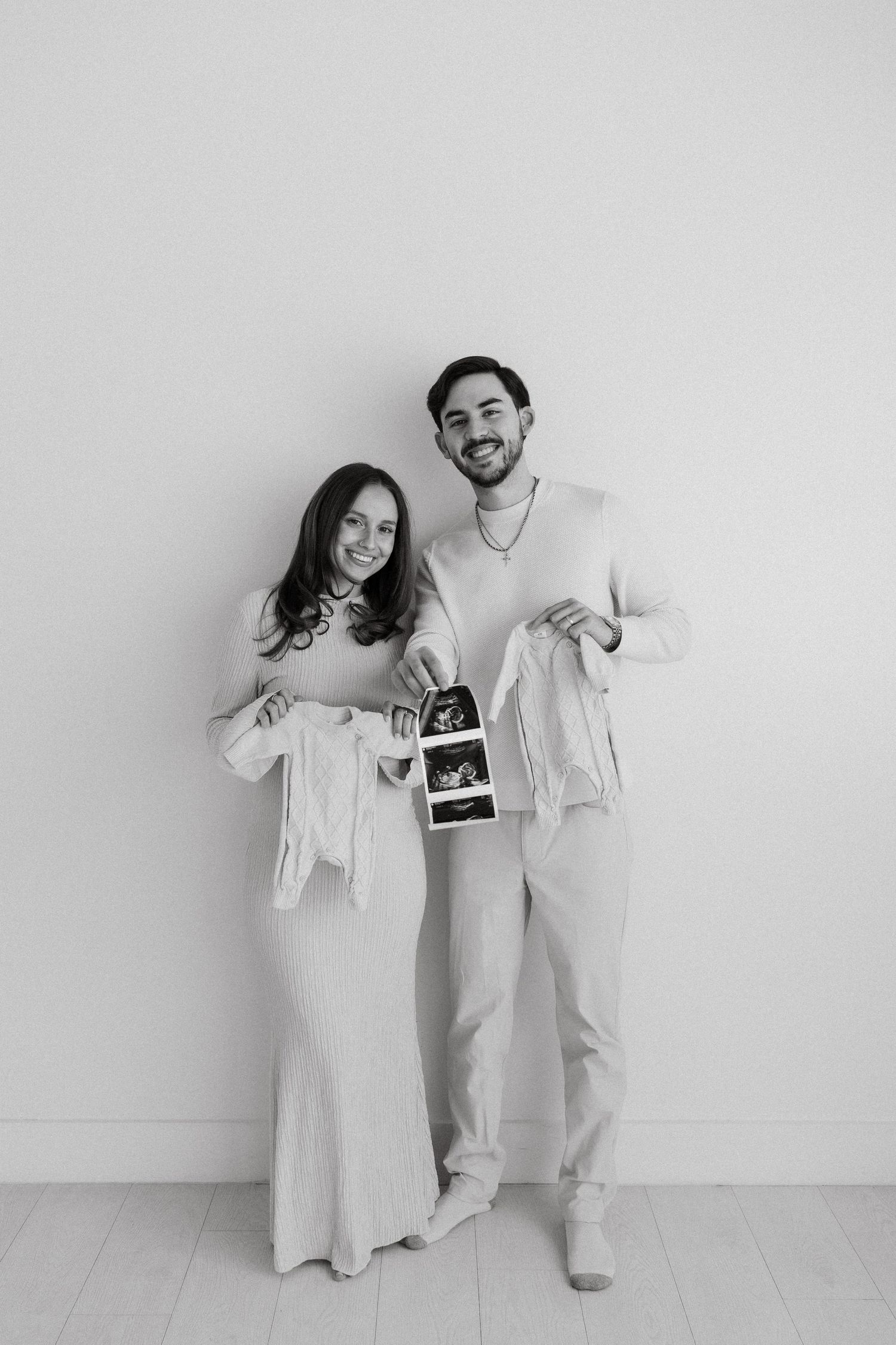 Happy couple holding ultrasound photos and baby clothes in black and white pregnancy announcement photo.