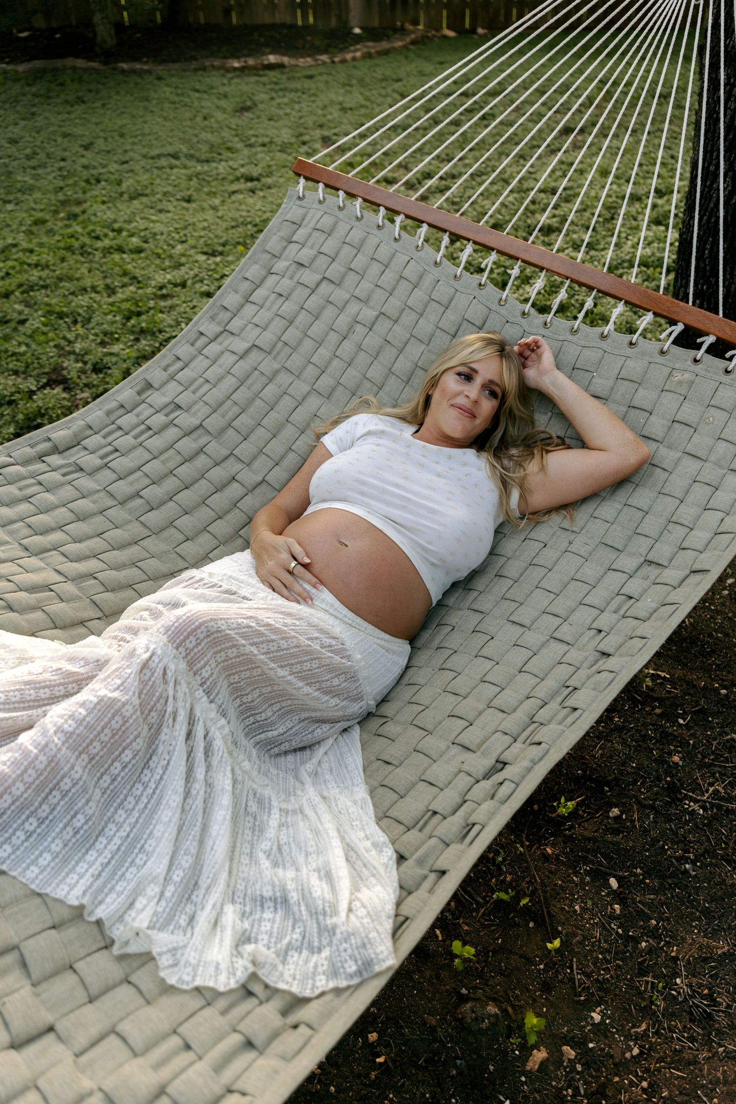 Pregnant woman in white two-piece outfit relaxing on a hammock during an outdoor maternity photo session.