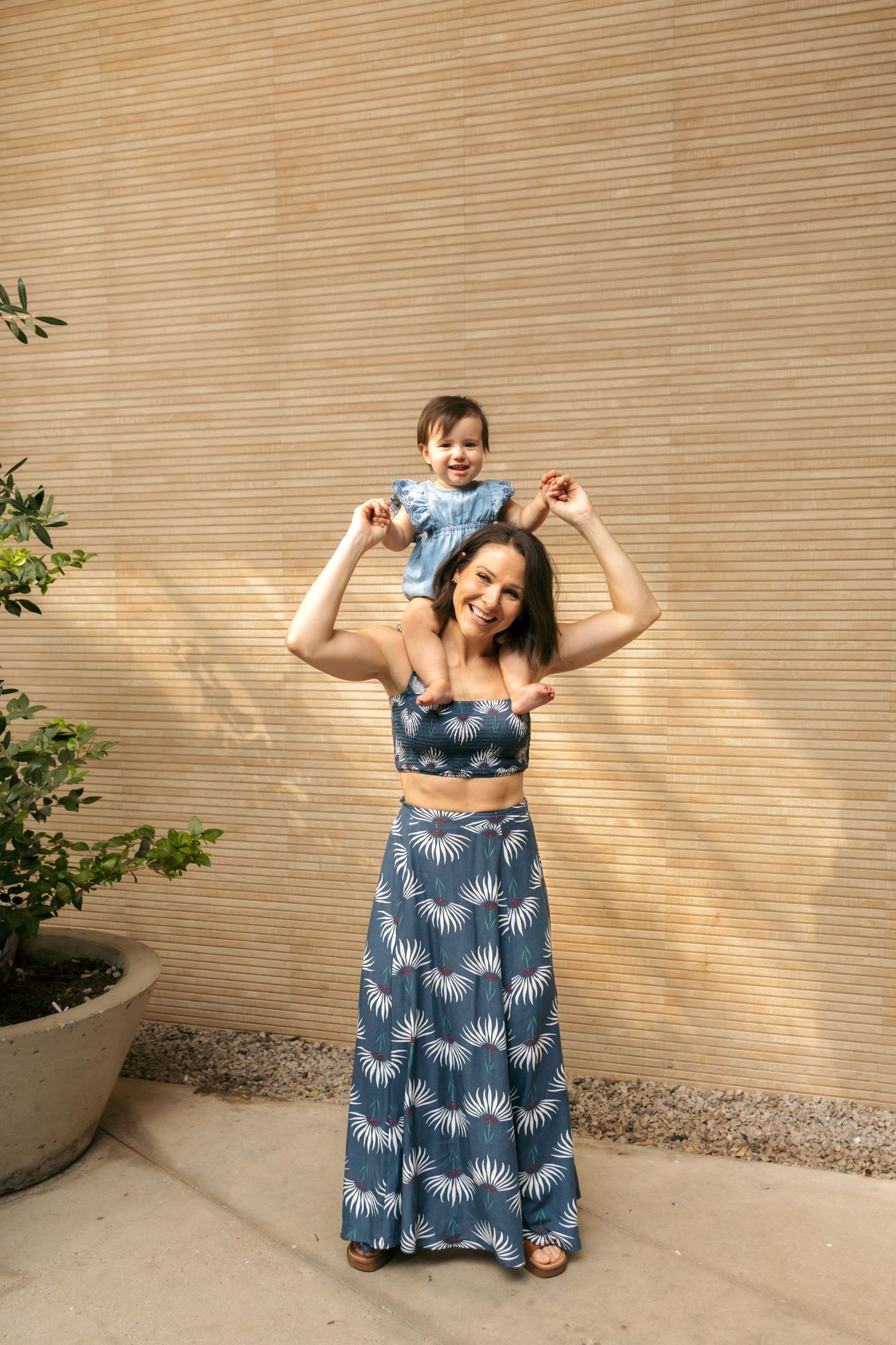 Smiling mother in blue floral matching set holding baby on shoulders against textured wall outdoors.