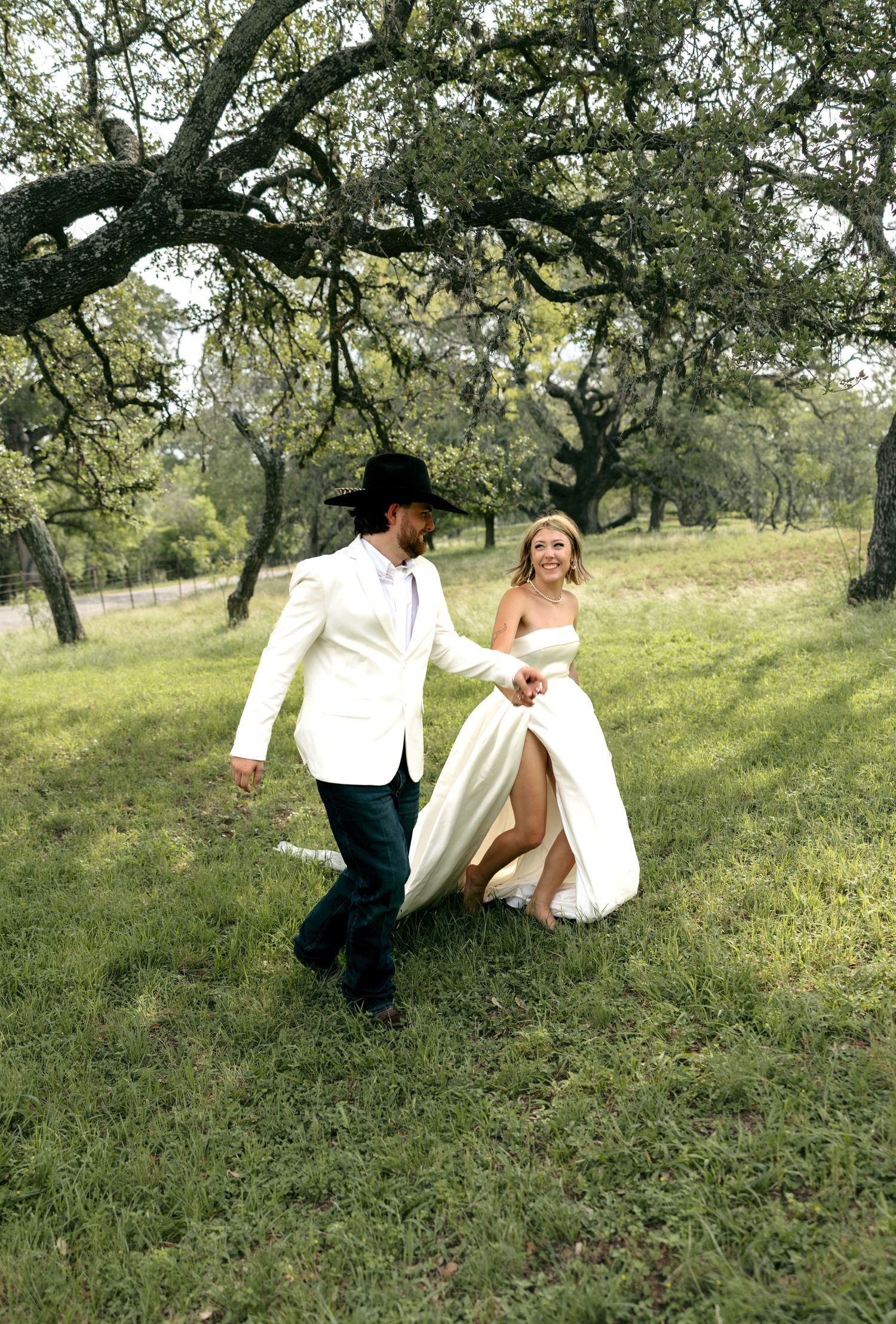 Bride and groom hold hands walking through green field under oak trees, groom in white jacket and black cowboy hat.