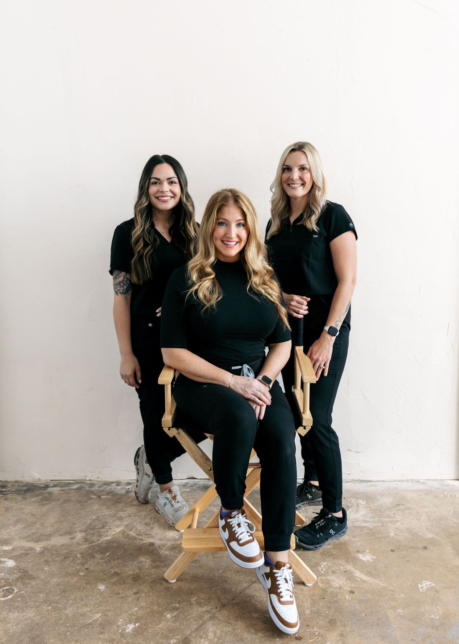 Three women in black scrubs posing together in a bright studio, one seated in directors chair.