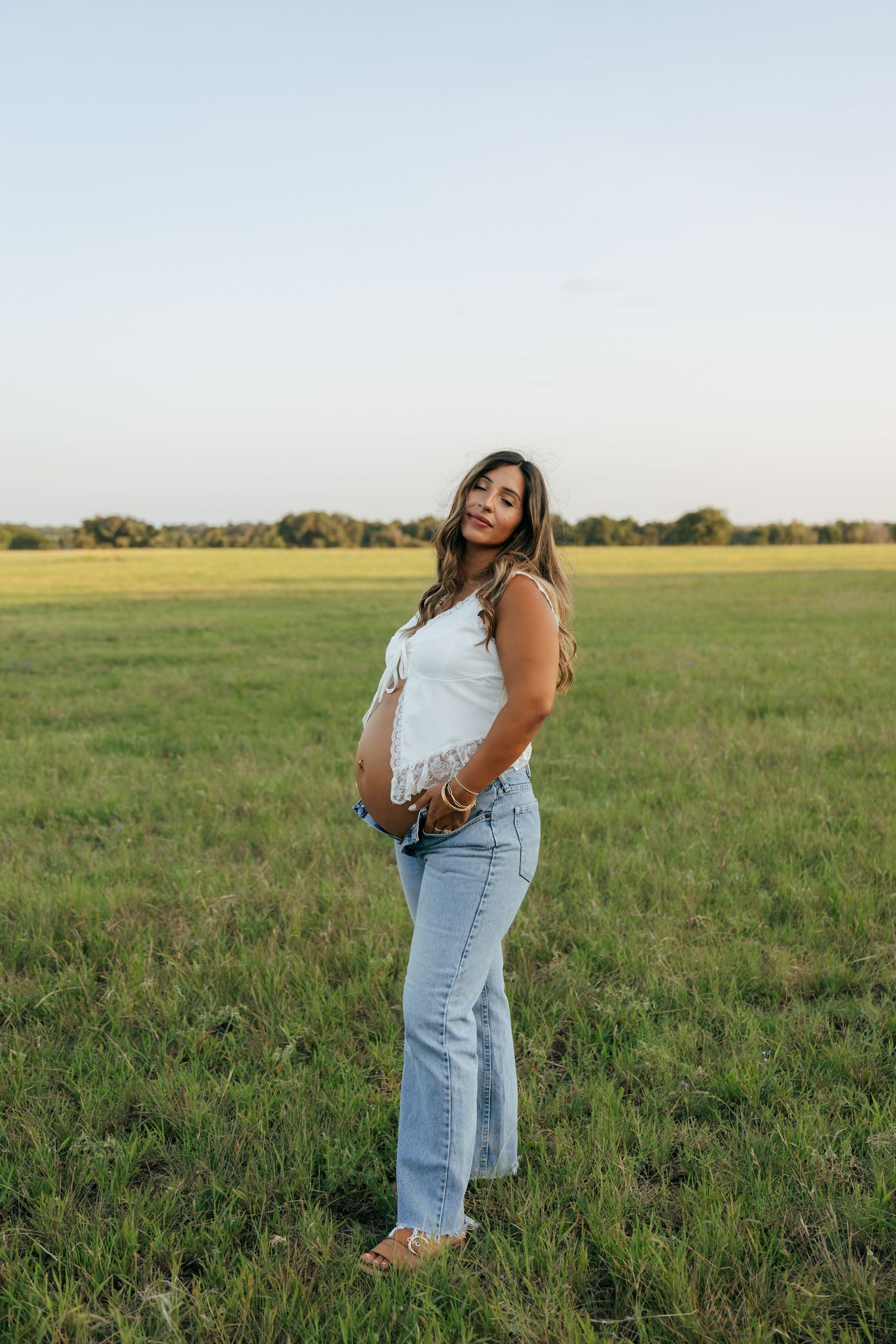 Pregnant woman in white top and jeans cradling baby bump in green field at golden hour.