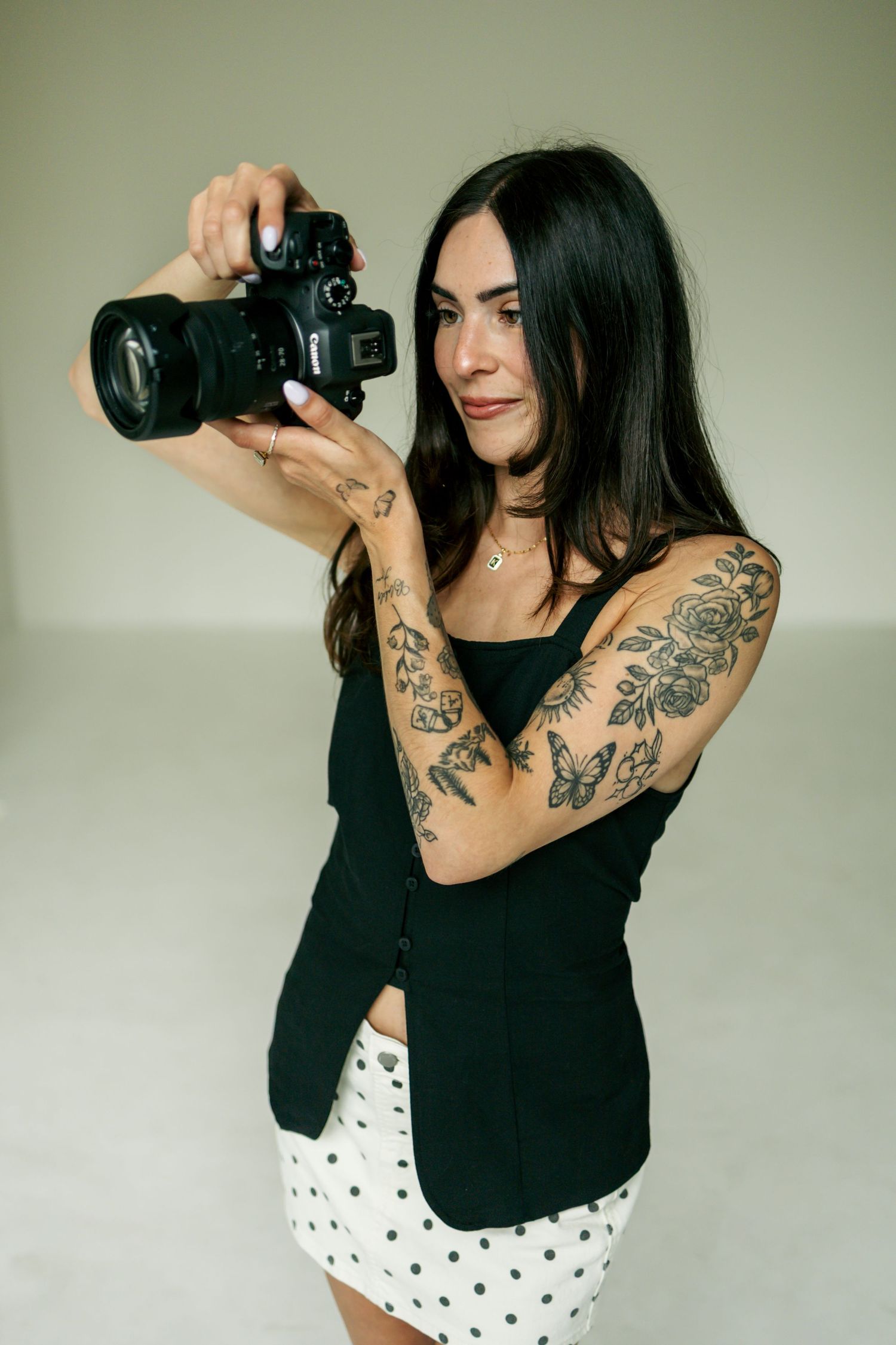 Tattooed female photographer in black blazer reviewing images on Canon DSLR camera in studio setting.