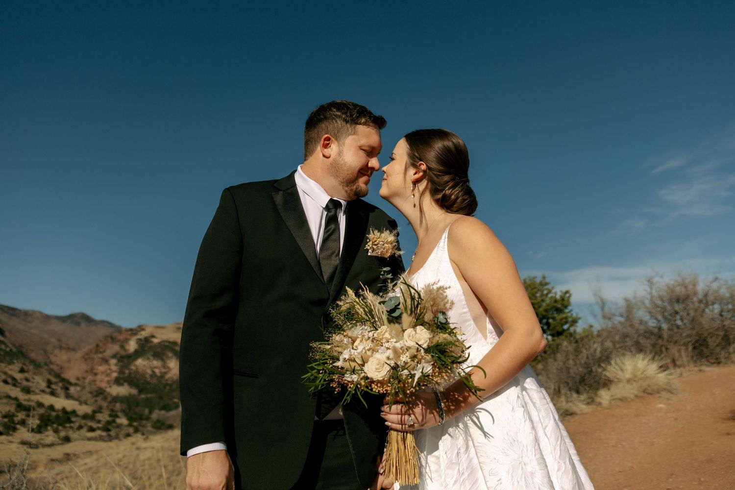 Bride holding boho bouquet and groom in black suit share a kiss in arid desert landscape under blue sky.