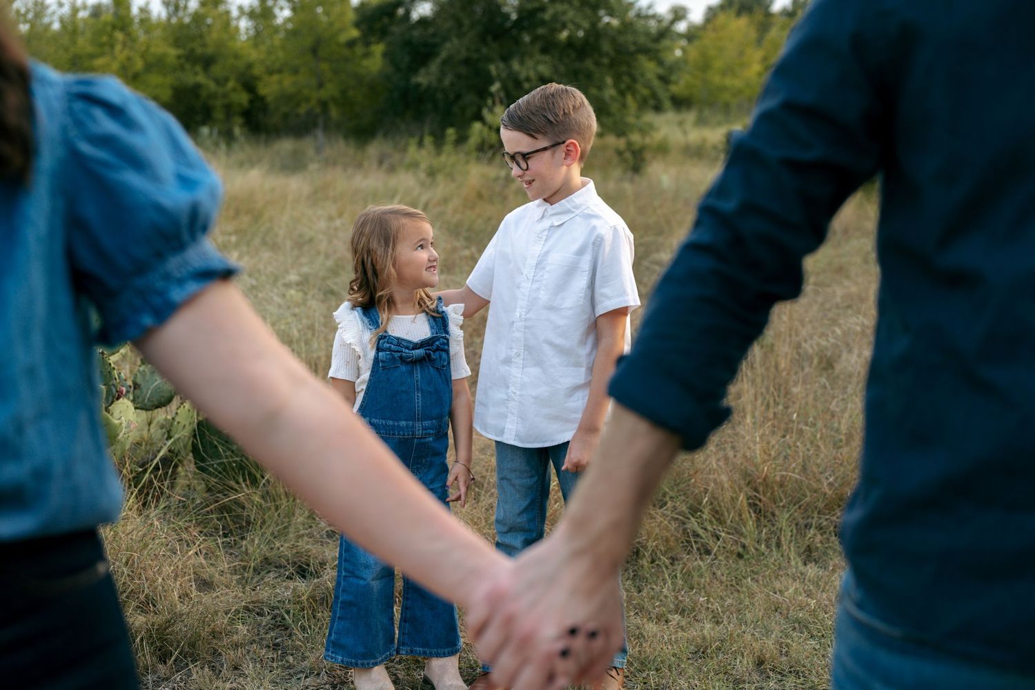 A boy and girl in matching denim outfits stand holding hands with parents in a grassy outdoor field.