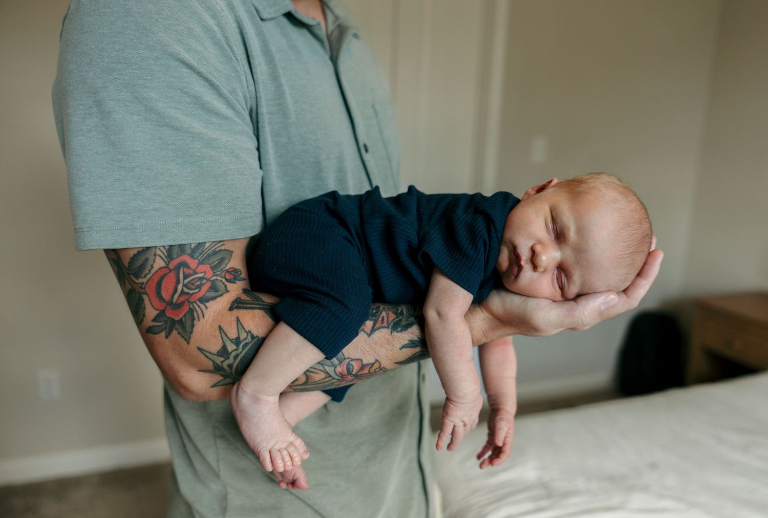 Tattooed father cradles sleeping newborn baby in one arm in a softly lit room.