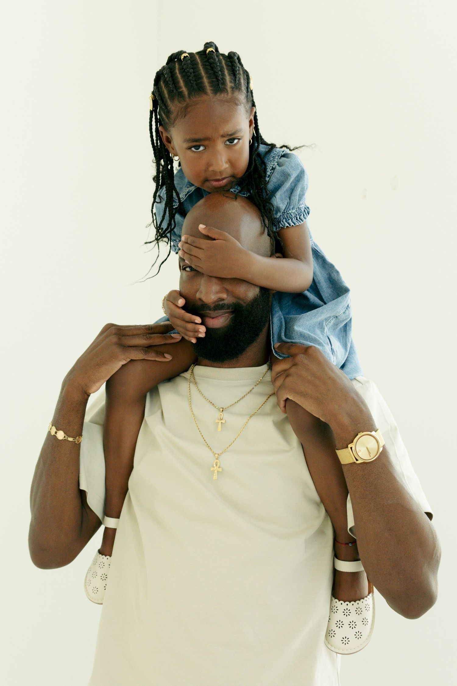 Father with braided-hair daughter sitting on his shoulders, wearing gold cross necklace, white background.