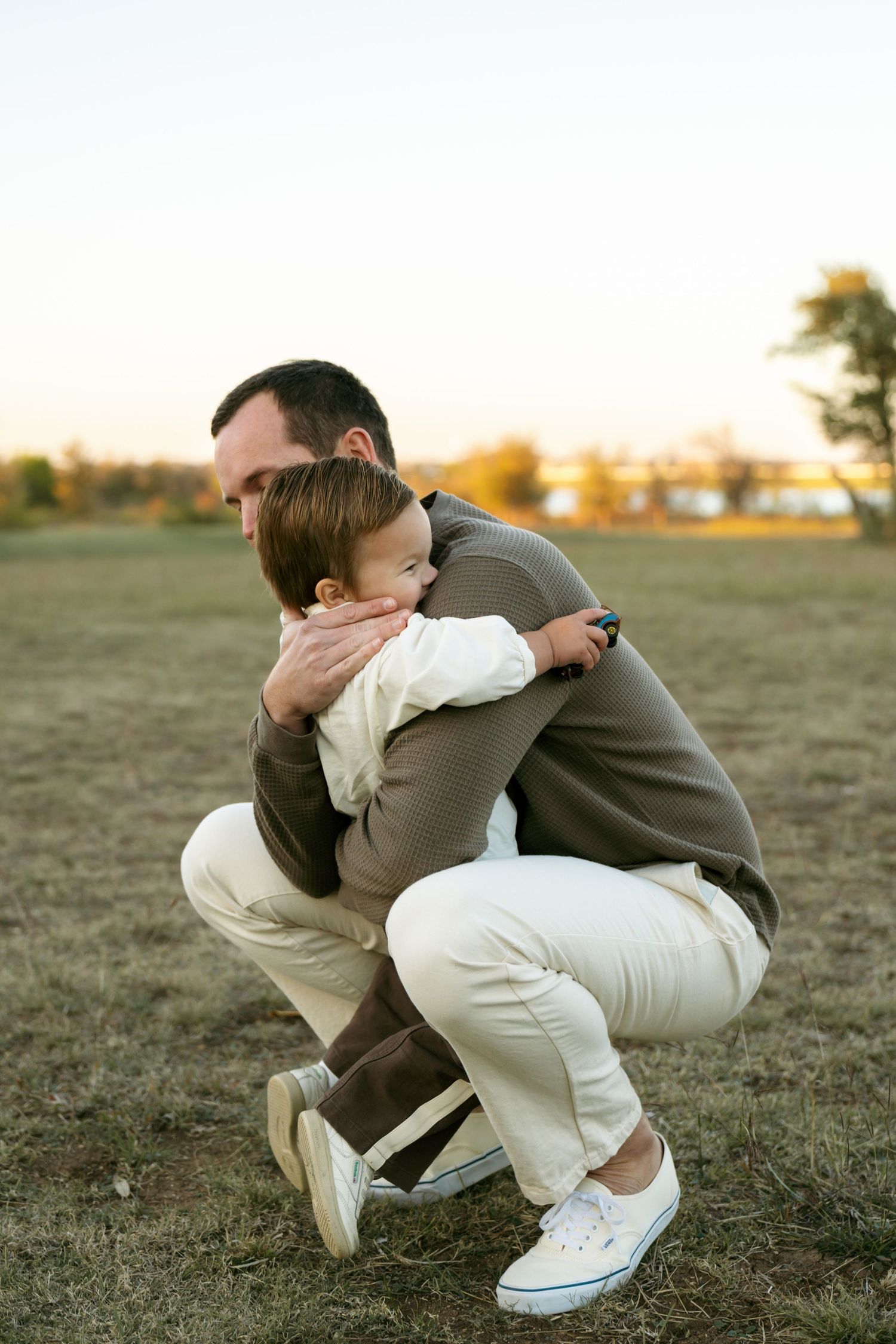 Father crouching down to embrace young toddler son in open field at sunset.