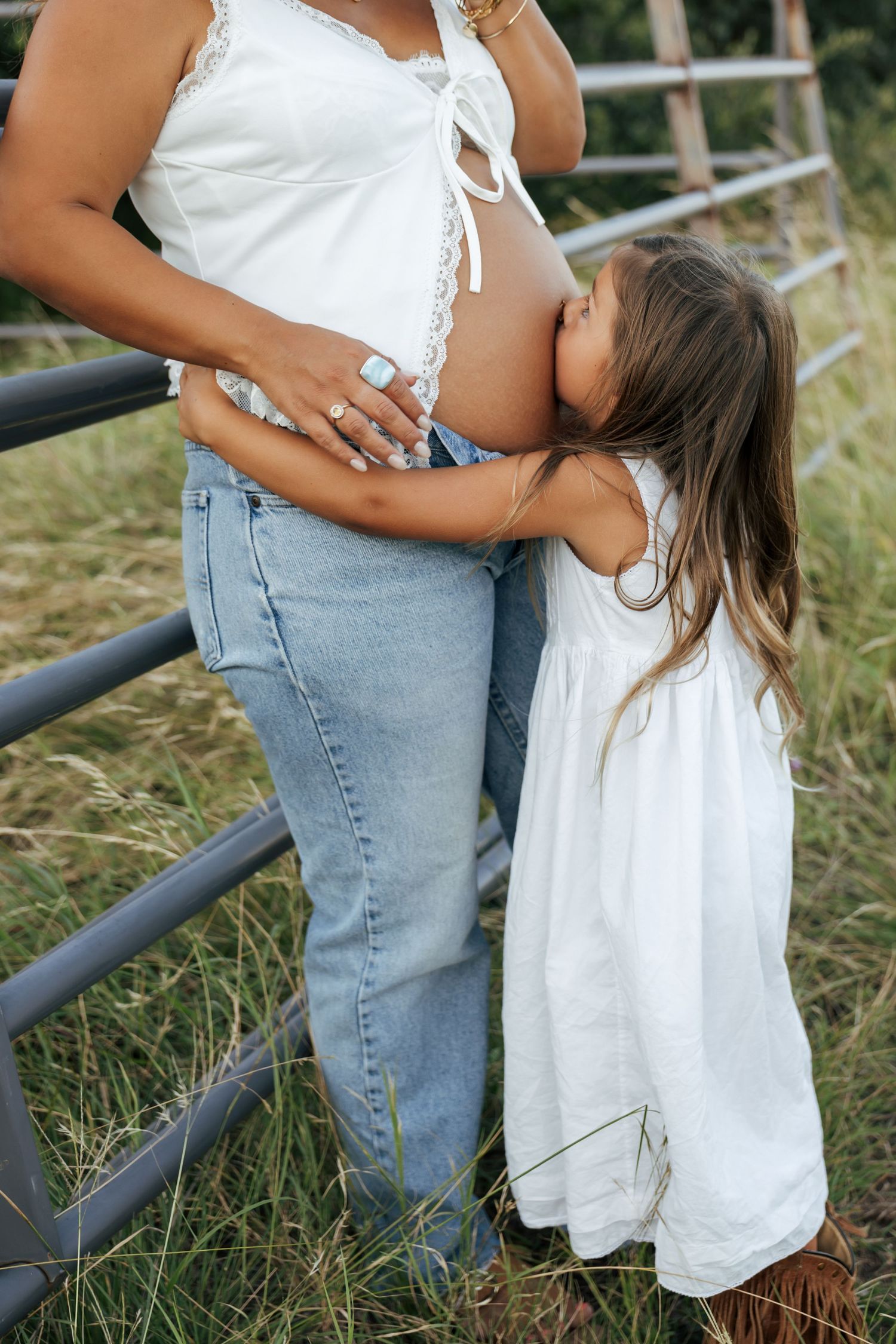 A young girl in a white dress kisses a pregnant woman's belly outdoors near a metal fence.