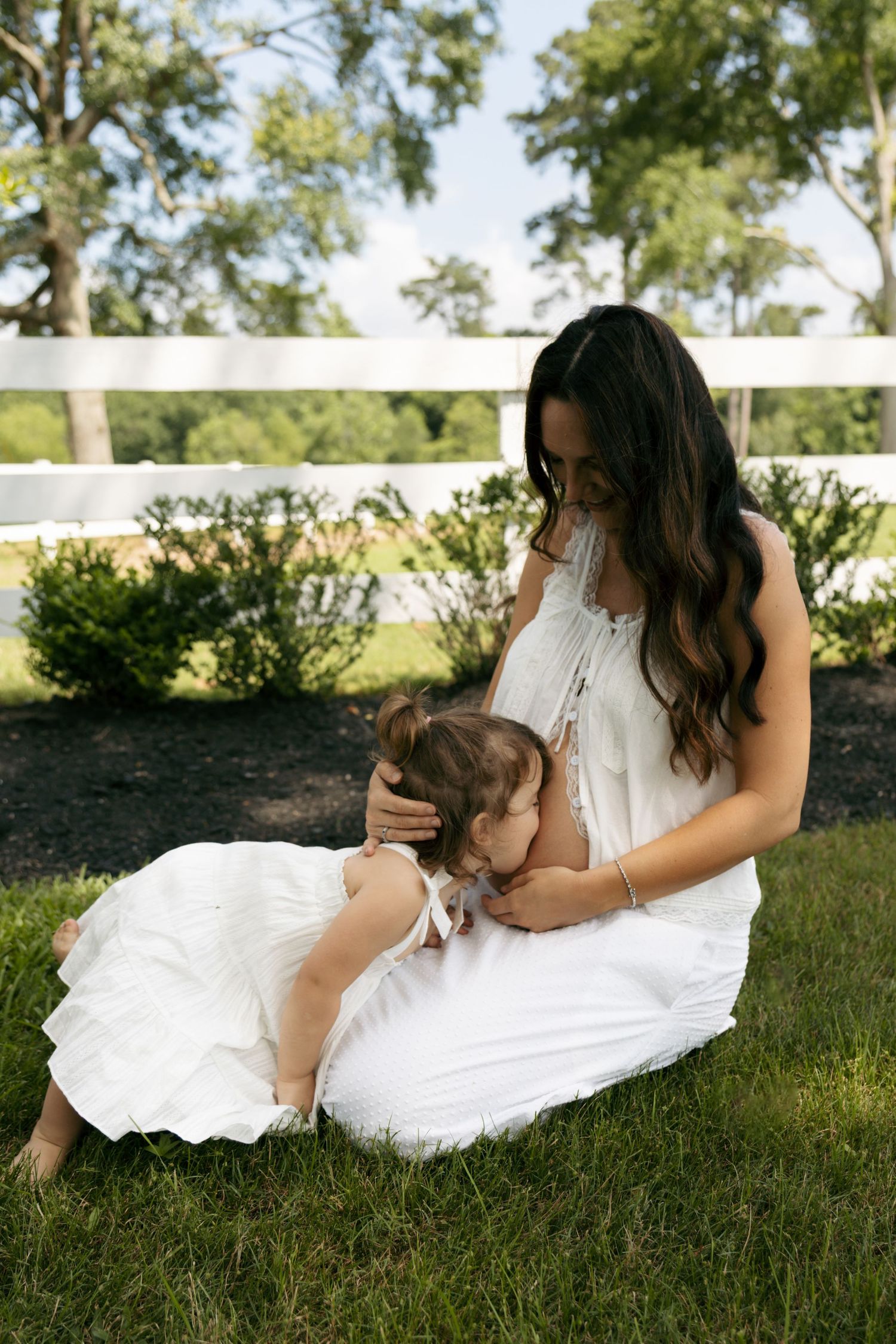 A dark-haired pregnant woman in white dress sits on grass hugging a toddler girl outdoors.