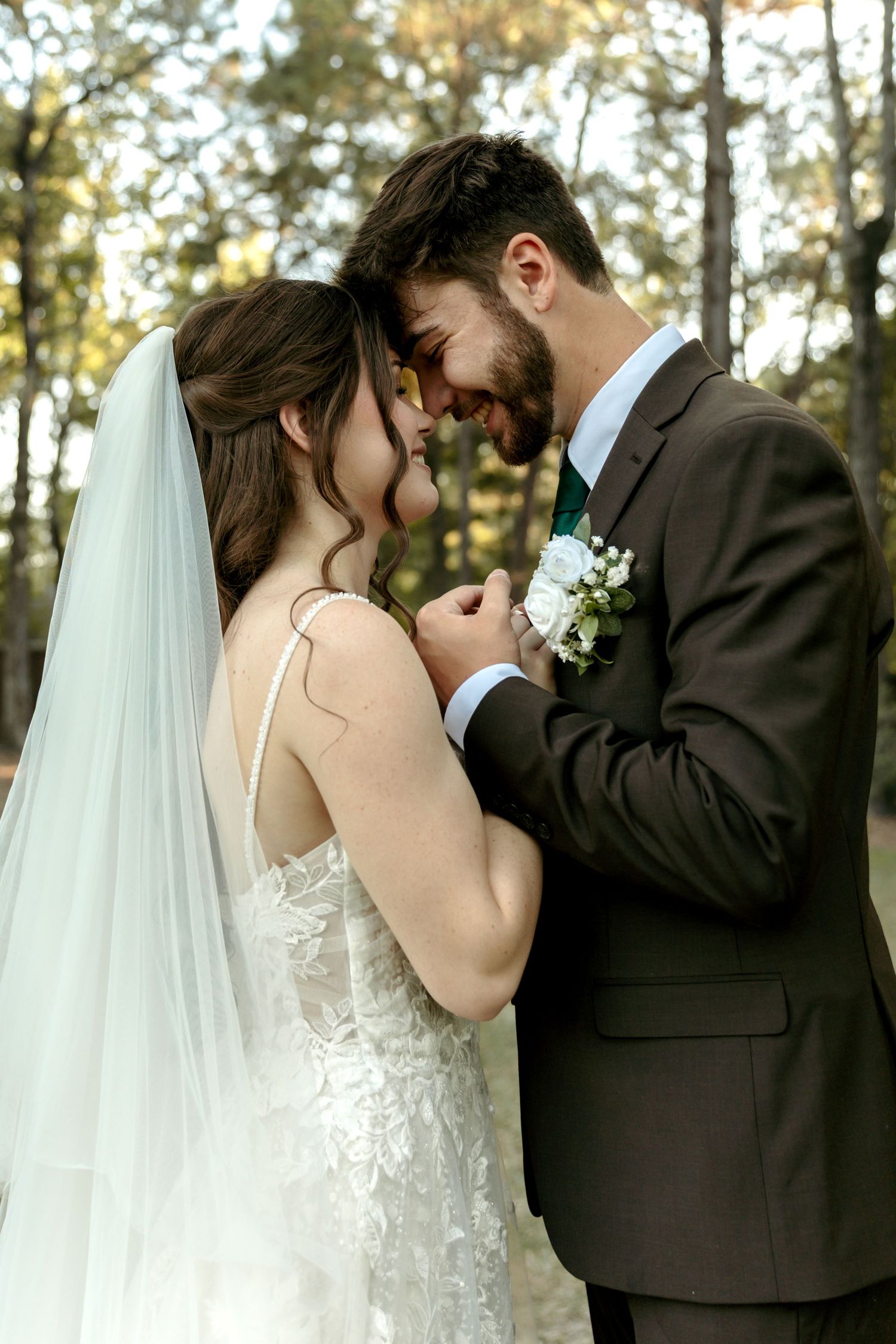 Bride and groom touching foreheads romantically in a sunlit forest setting.