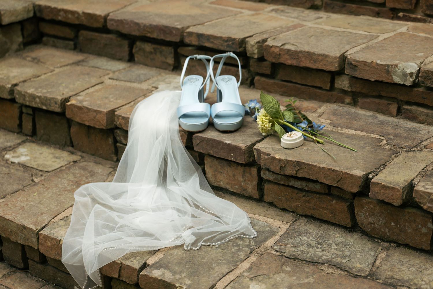 Light blue bridal heels and floral bouquet resting on rustic stone steps beside a flowing white wedding veil.