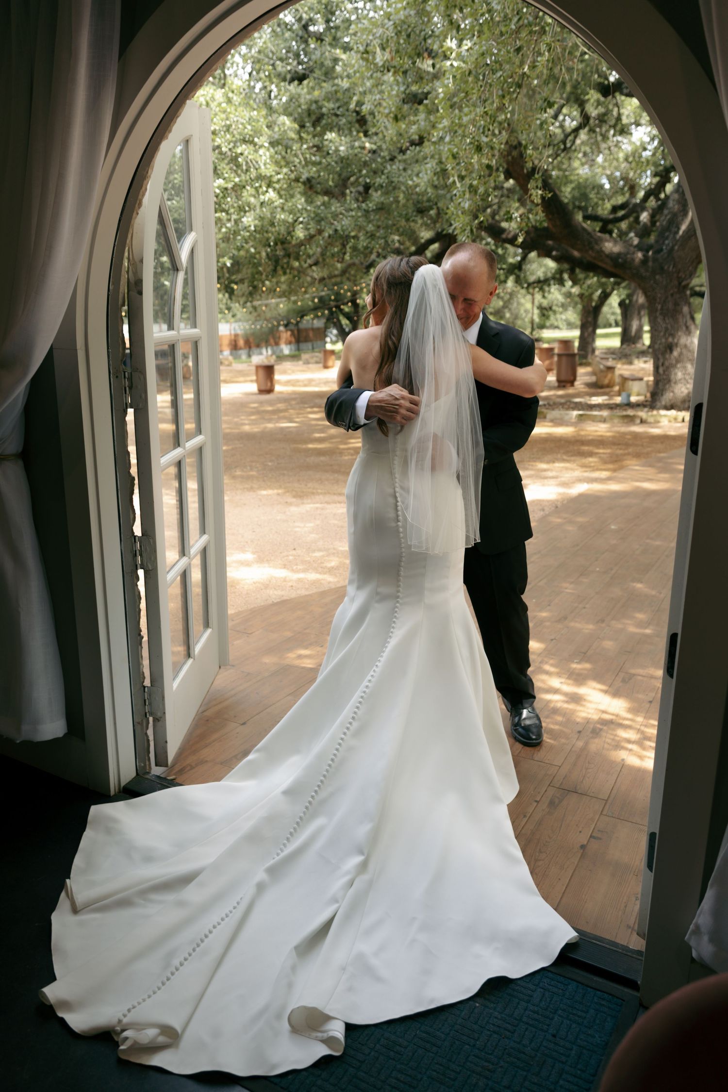 A bride in a mermaid gown and groom share a dance framed by an arched doorway opening to a tree-lined path.
