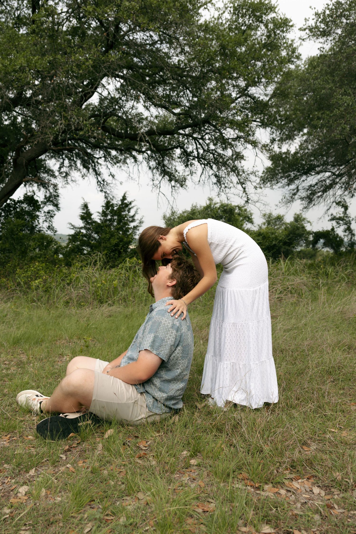 A woman in a white dress leans down to kiss a man sitting on grass beneath a large tree outdoors.