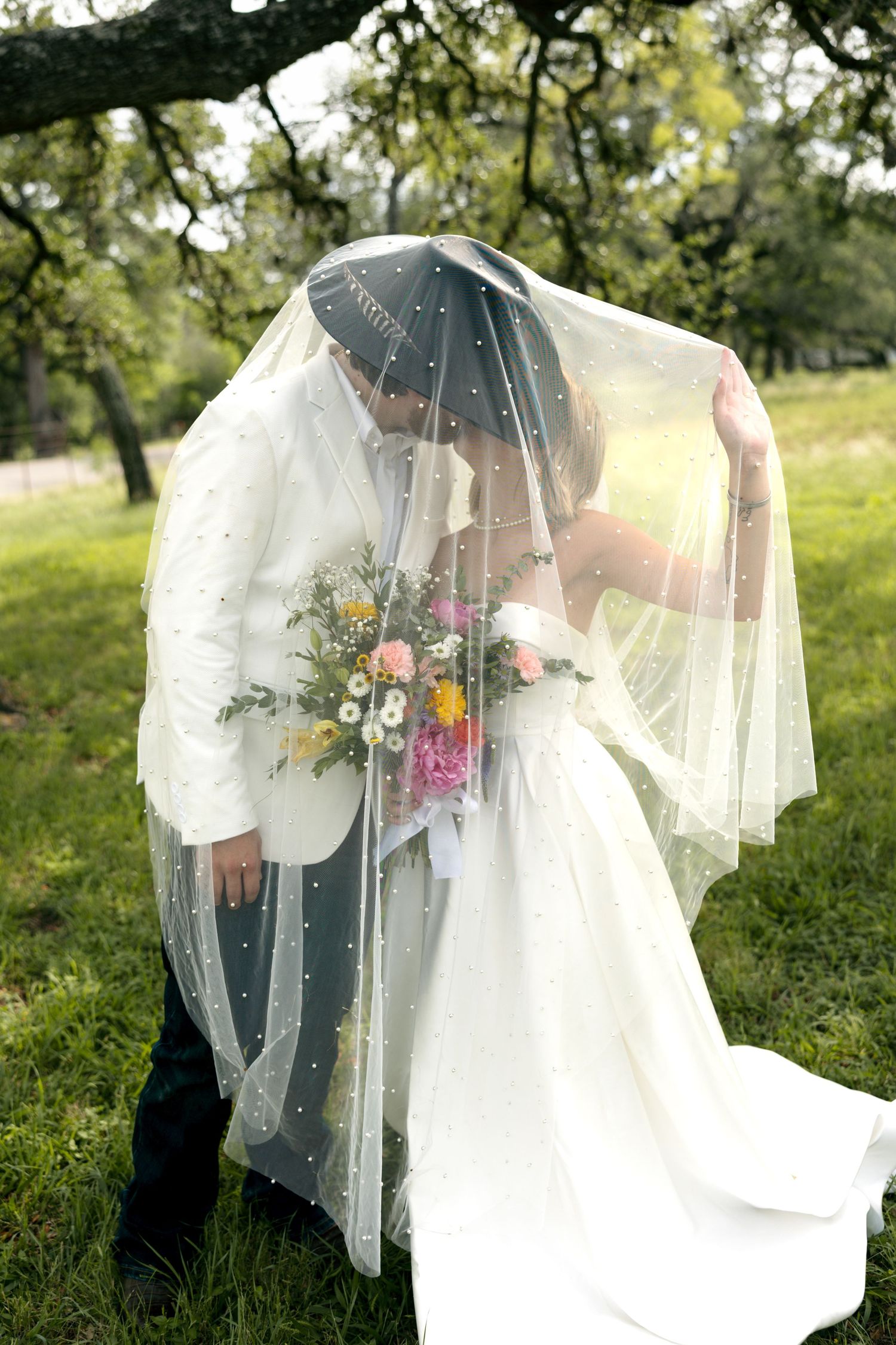 Bride and groom share a kiss hidden under a polka dot veil in a lush green outdoor setting.