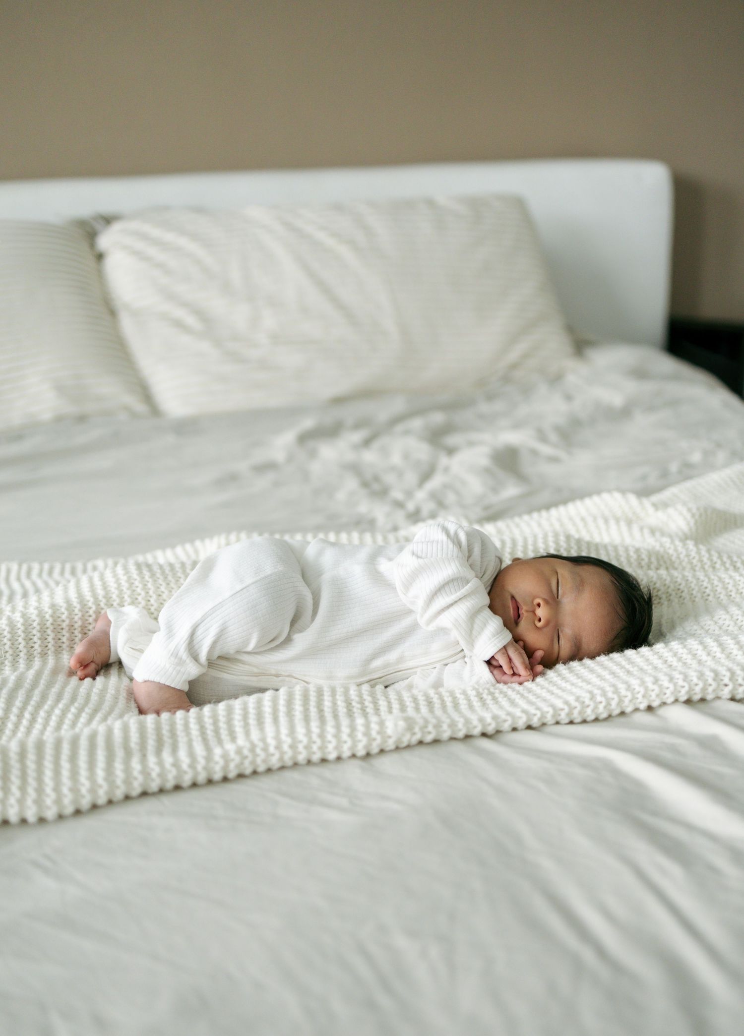 Newborn baby sleeping peacefully in white onesie on a white textured blanket on a bed.