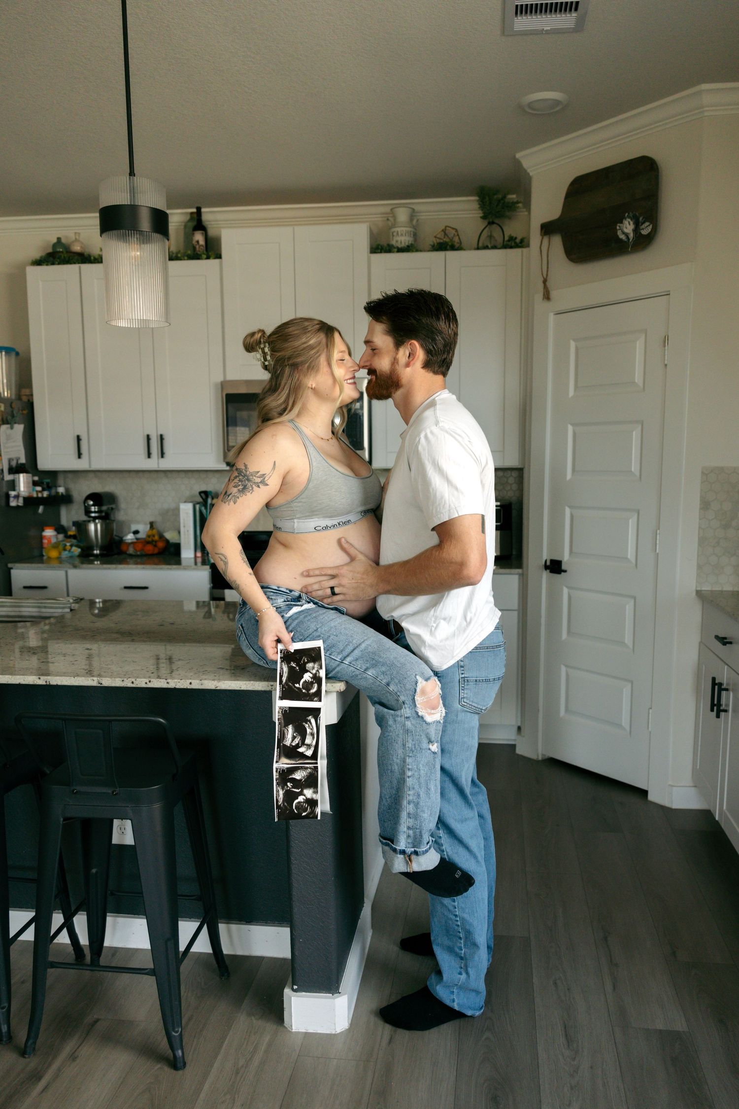 A pregnant couple shares a romantic moment in a modern kitchen while holding ultrasound photos.