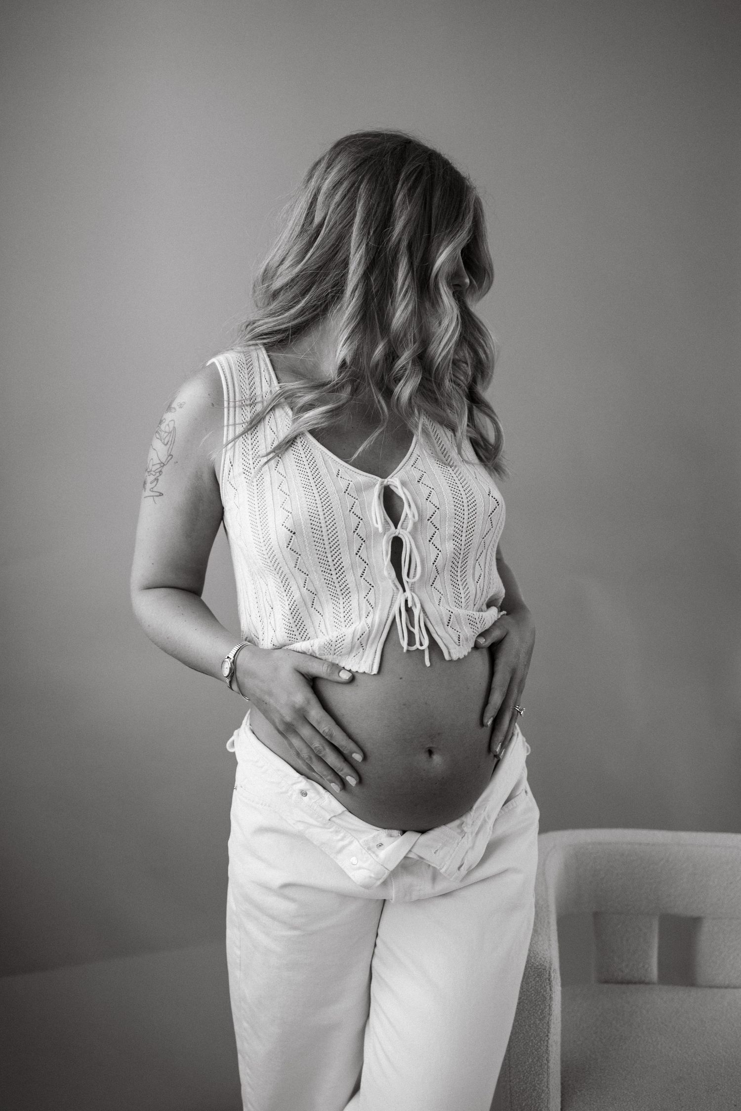 Black and white photo of pregnant woman in white crop top holding baby bump indoors.