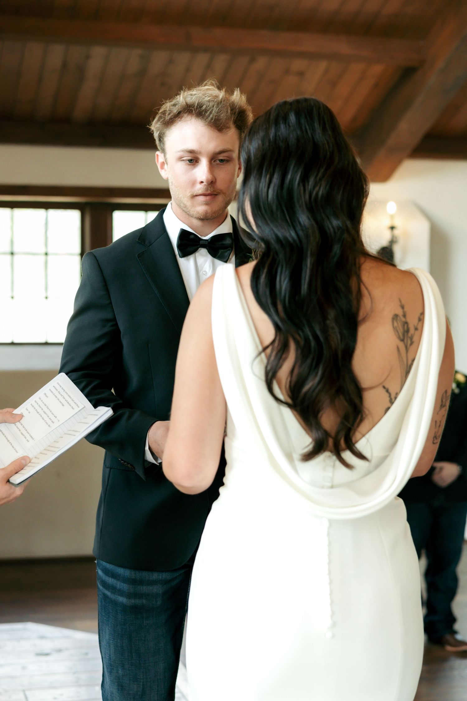 Groom in black tuxedo reads wedding vows to bride in backless white gown with floral tattoo during indoor ceremony.
