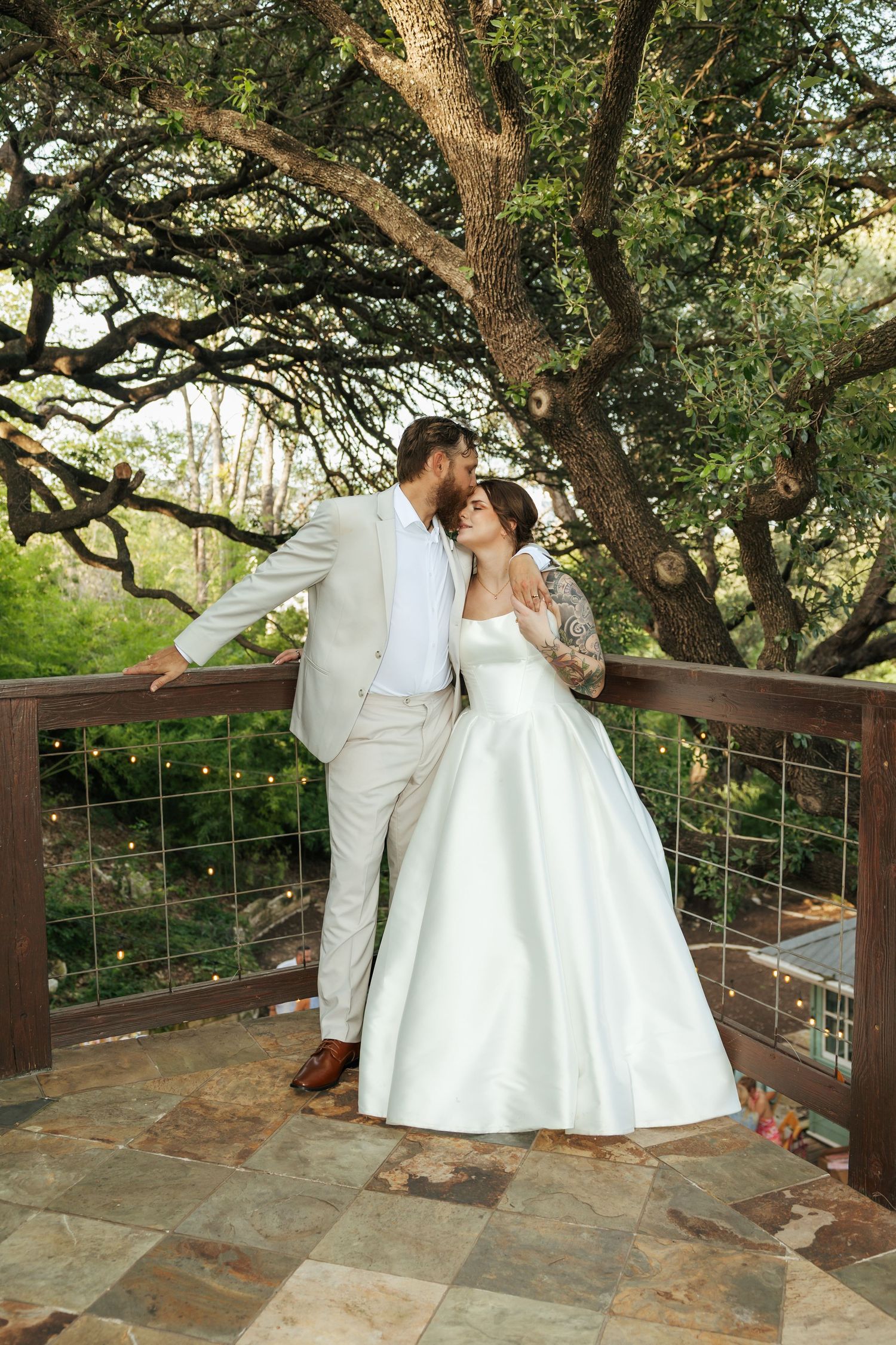 Bride and groom sharing a romantic moment on a stone patio surrounded by large oak trees.