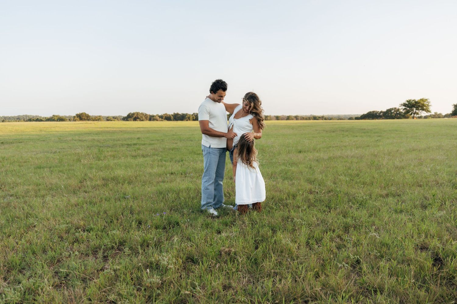 Family of three standing together in a vast green field during a golden hour session.