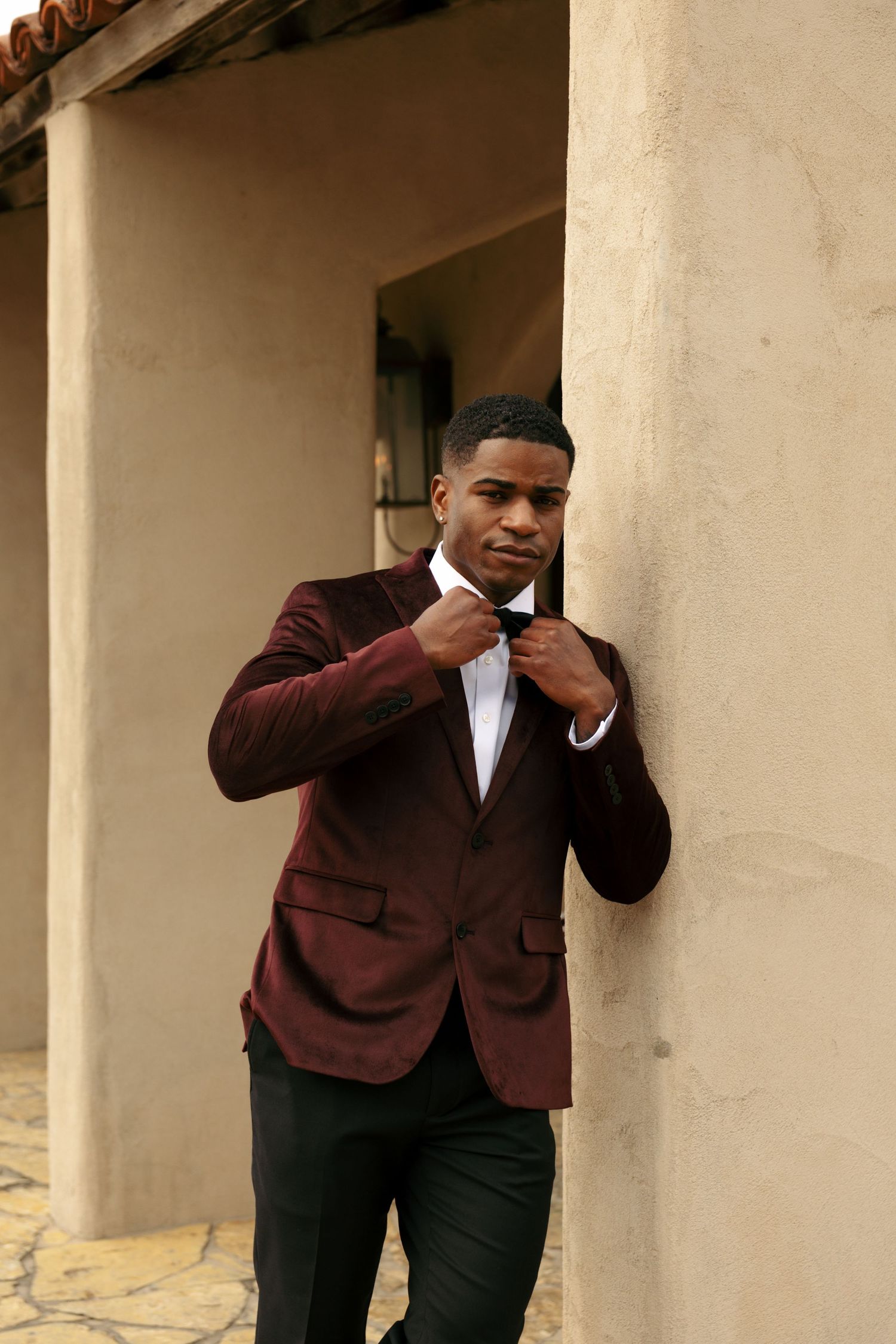 Stylish young man in burgundy velvet blazer and white shirt leans against beige stucco wall outdoors.