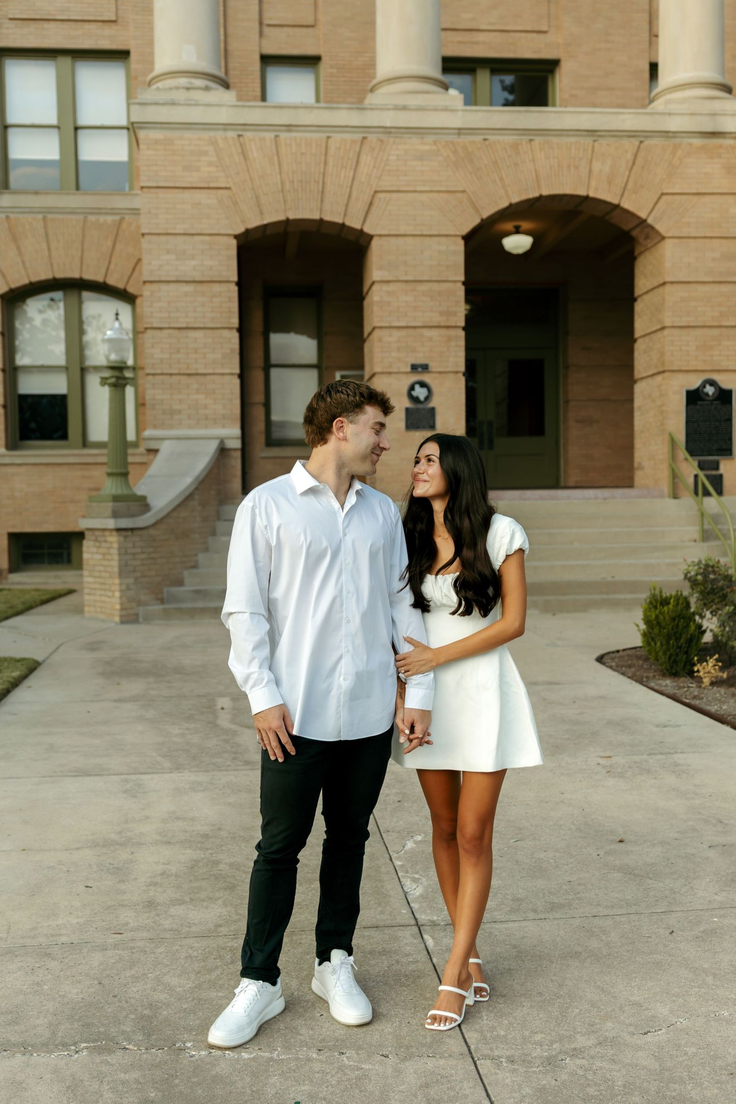 Couple in matching white outfits holding hands and smiling at each other in front of a brick building.