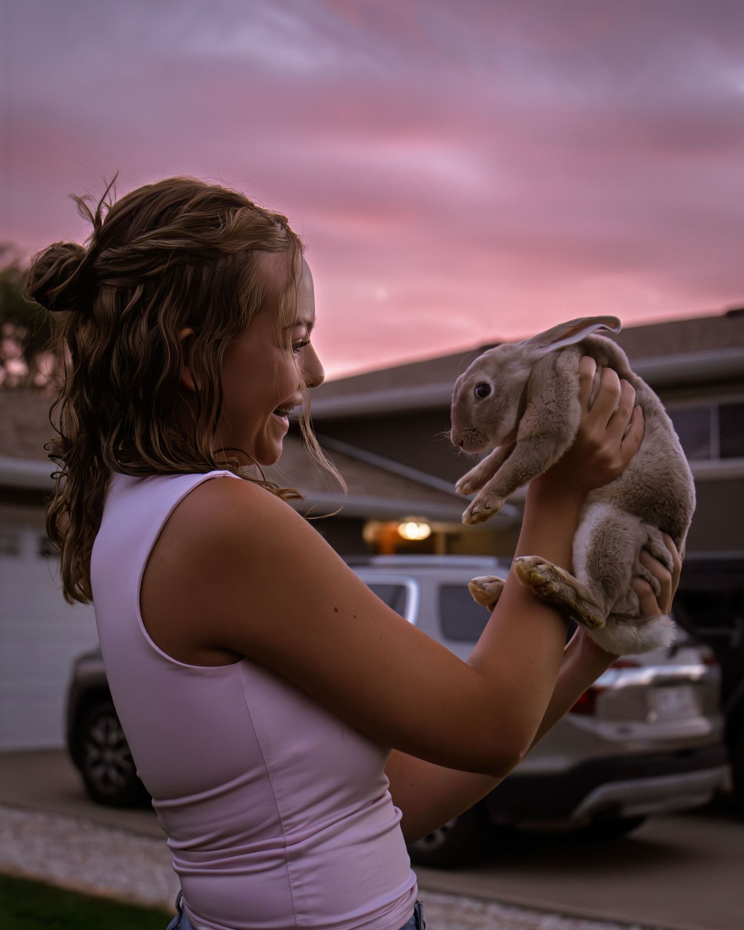 Smiling young girl holding a rabbit against a vibrant pink sunset sky in a driveway.