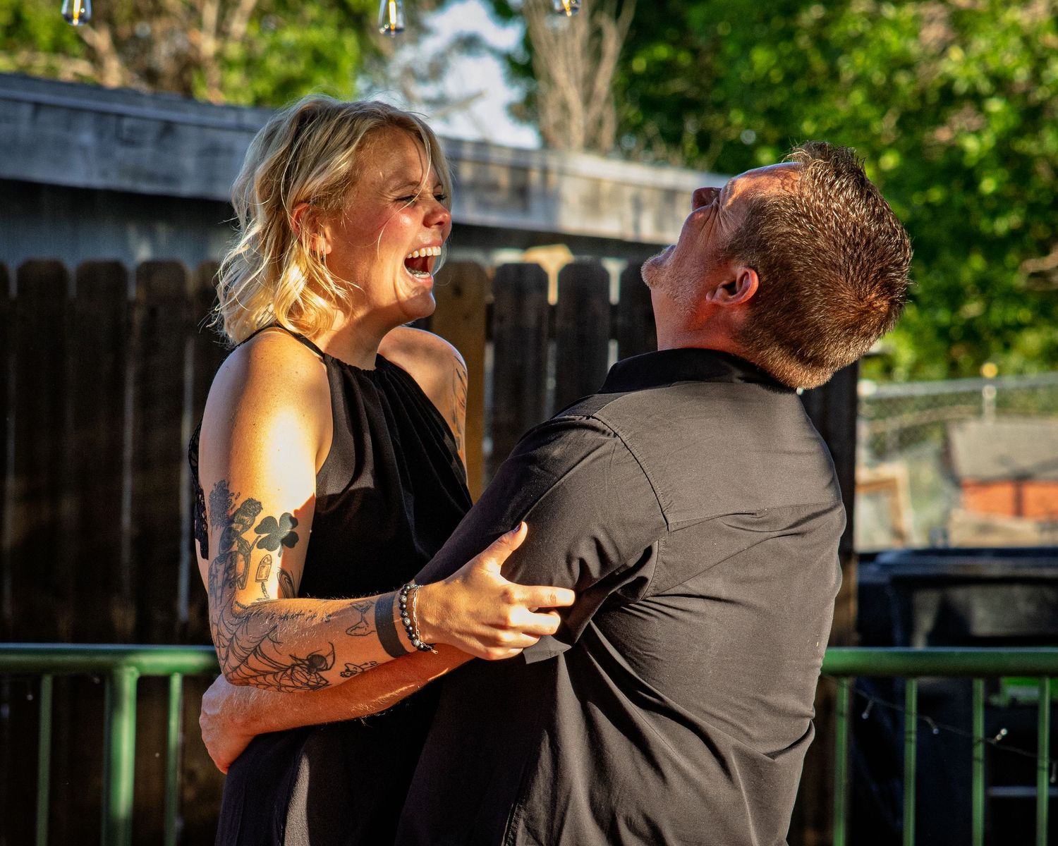 A tattooed woman in a black dress laughs joyfully while dancing with a man at an outdoor party.