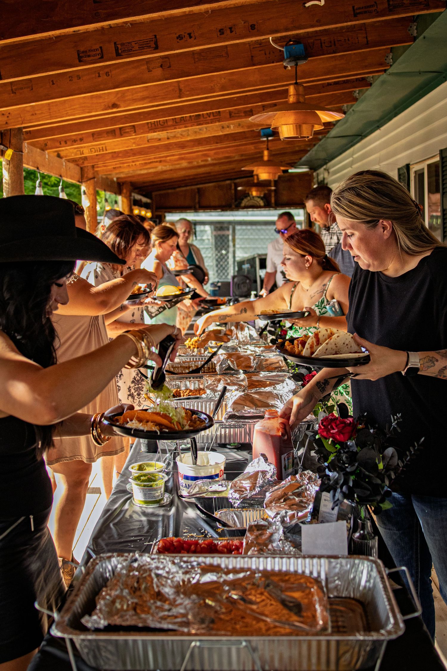 Guests serving themselves from a buffet of food trays under warm wooden ceiling lights at an outdoor covered patio event.