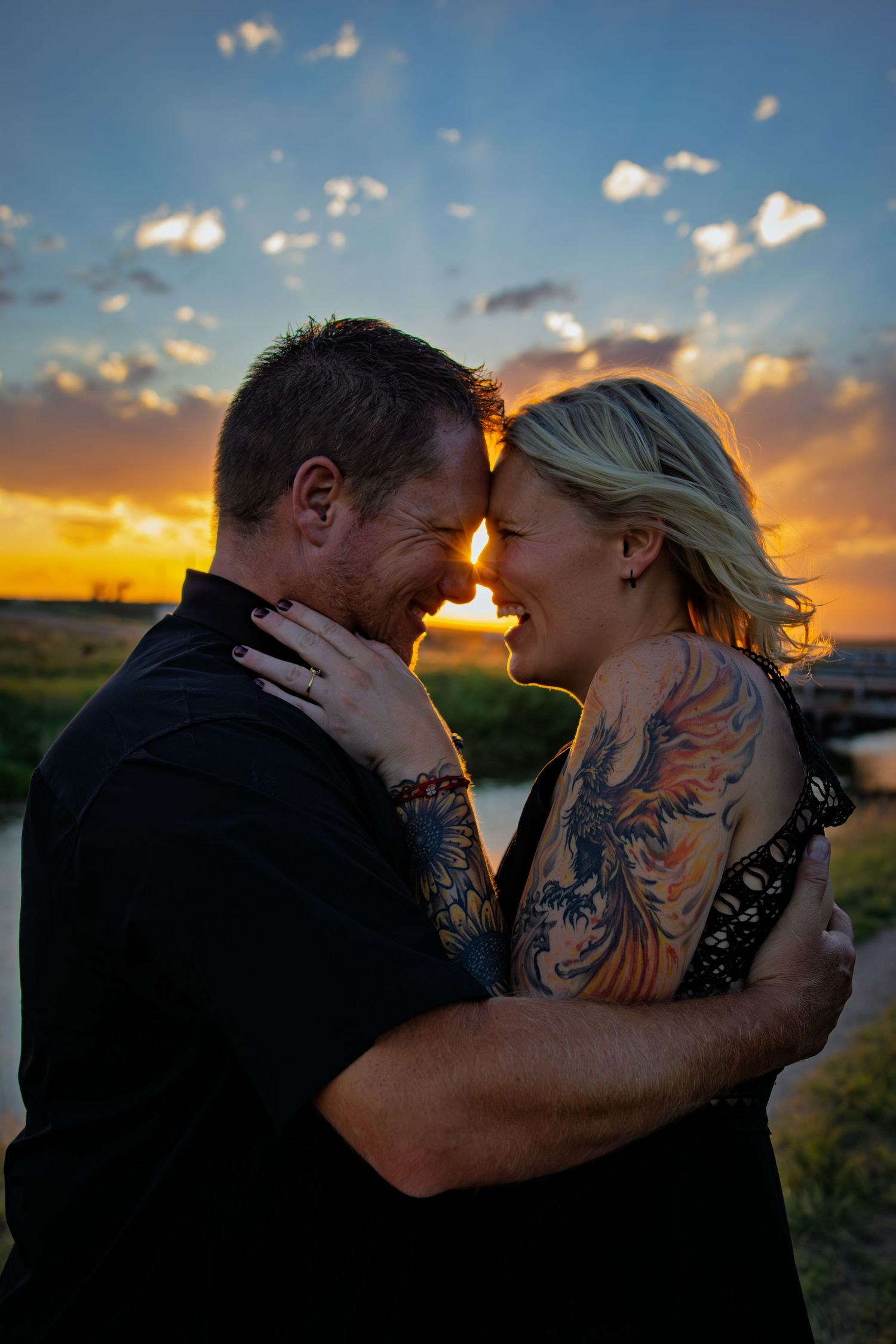 Tattooed couple embracing and smiling at sunset with colorful clouds behind them.