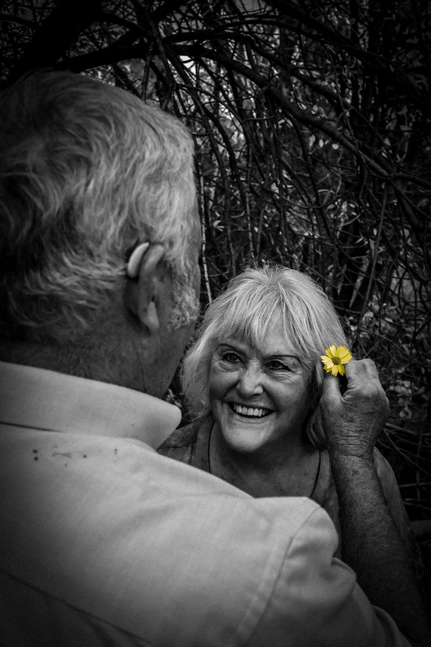 A black and white photo of an elderly man placing a yellow flower in a smiling womans hair.