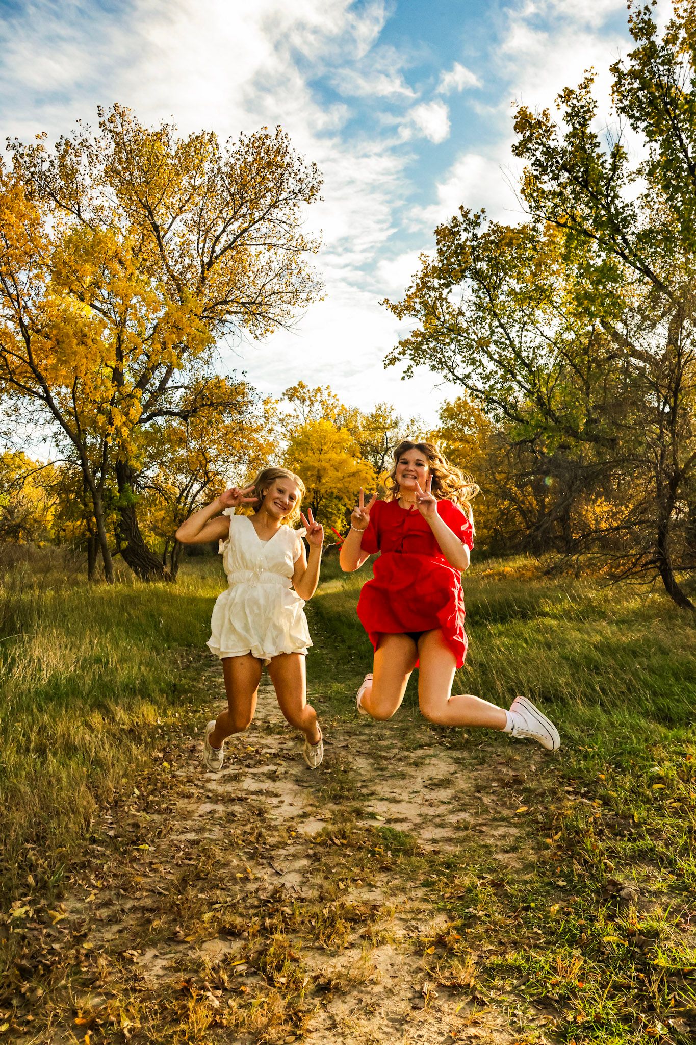 Two girls in white and red dresses jump joyfully on a dirt path surrounded by fall foliage.