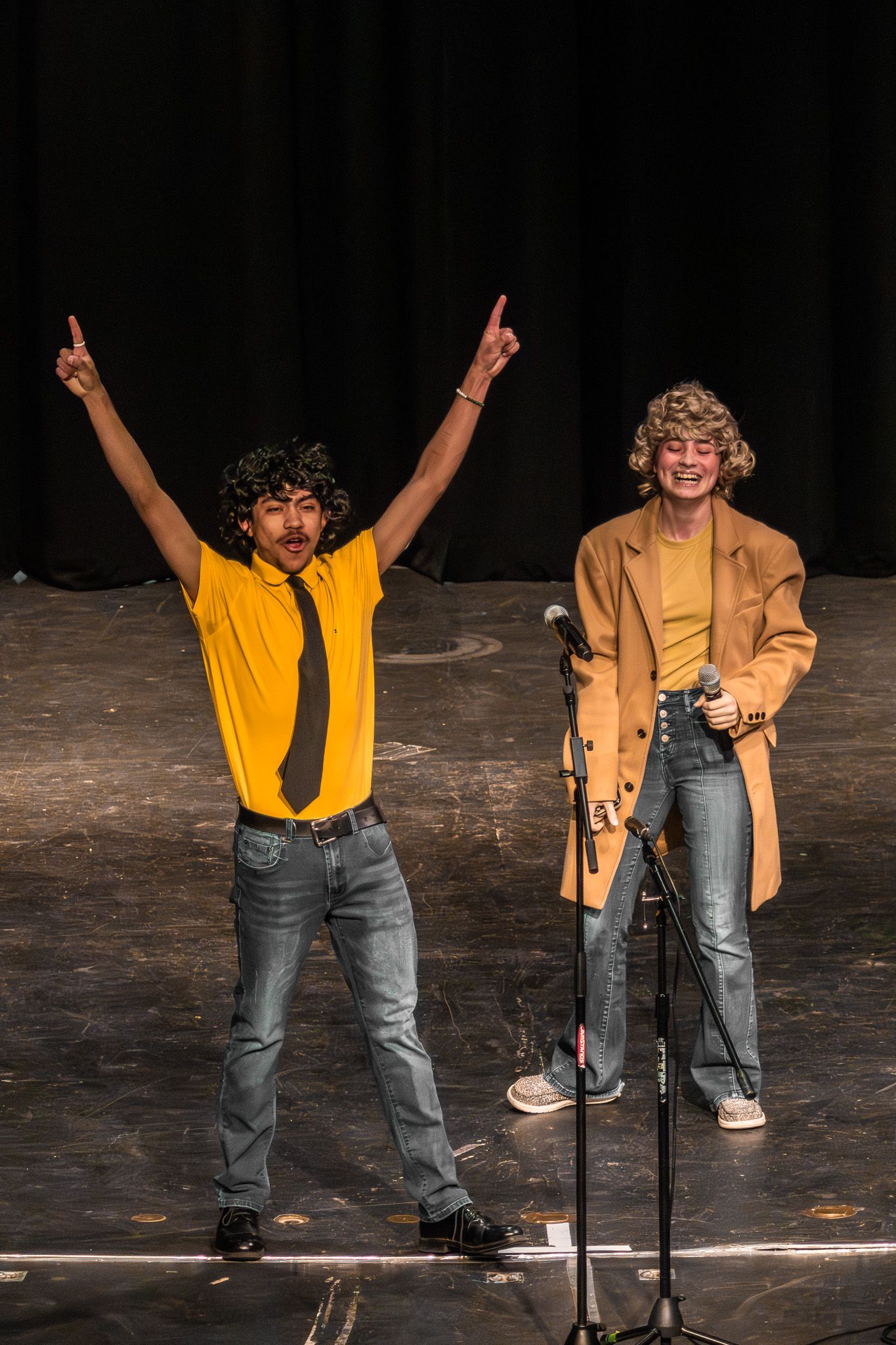 Two male performers celebrating on stage, one raising arms in yellow shirt, at live talent show.