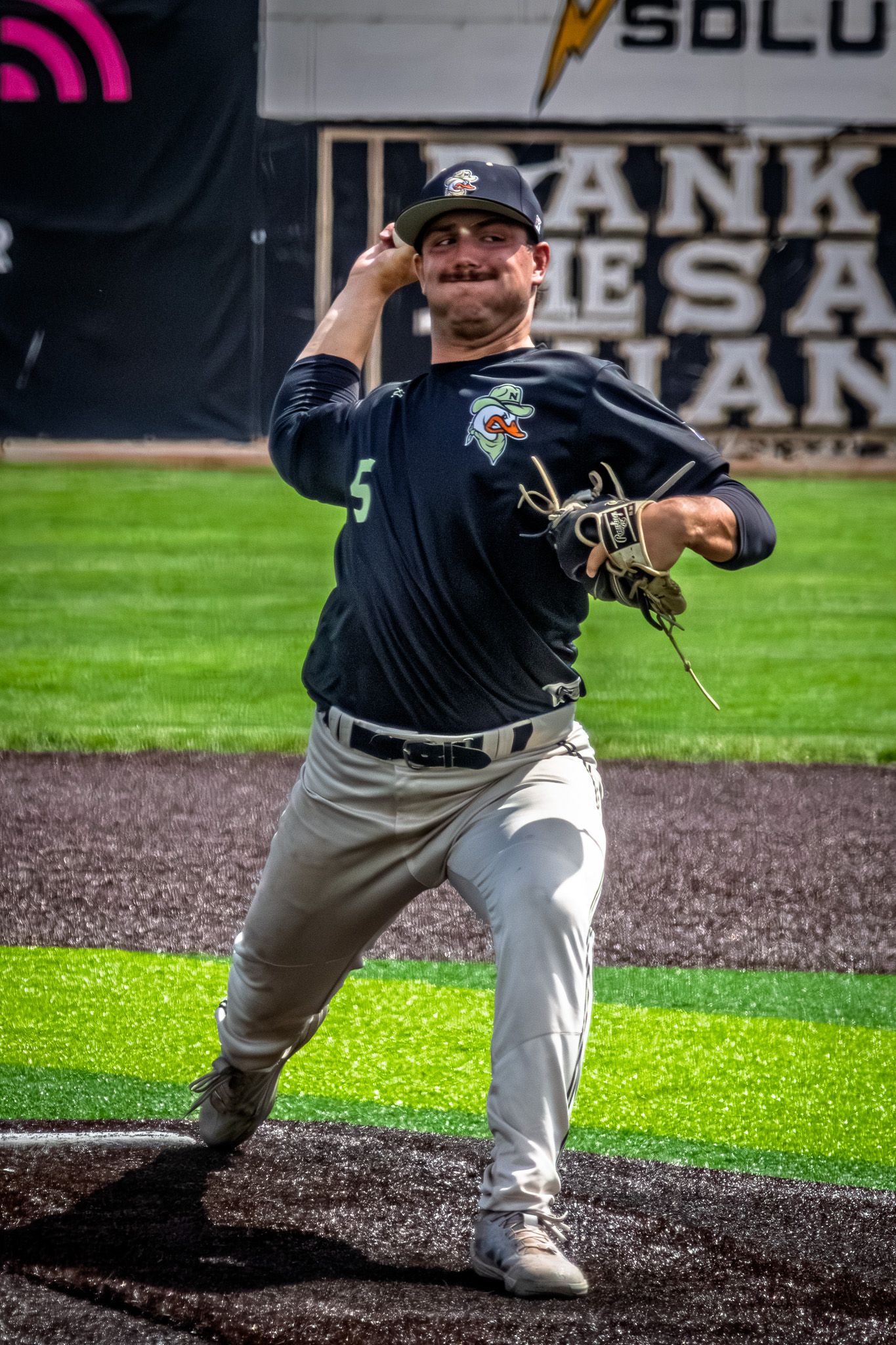 A smiling baseball pitcher in dark uniform winds up to throw during a game at an outdoor stadium.